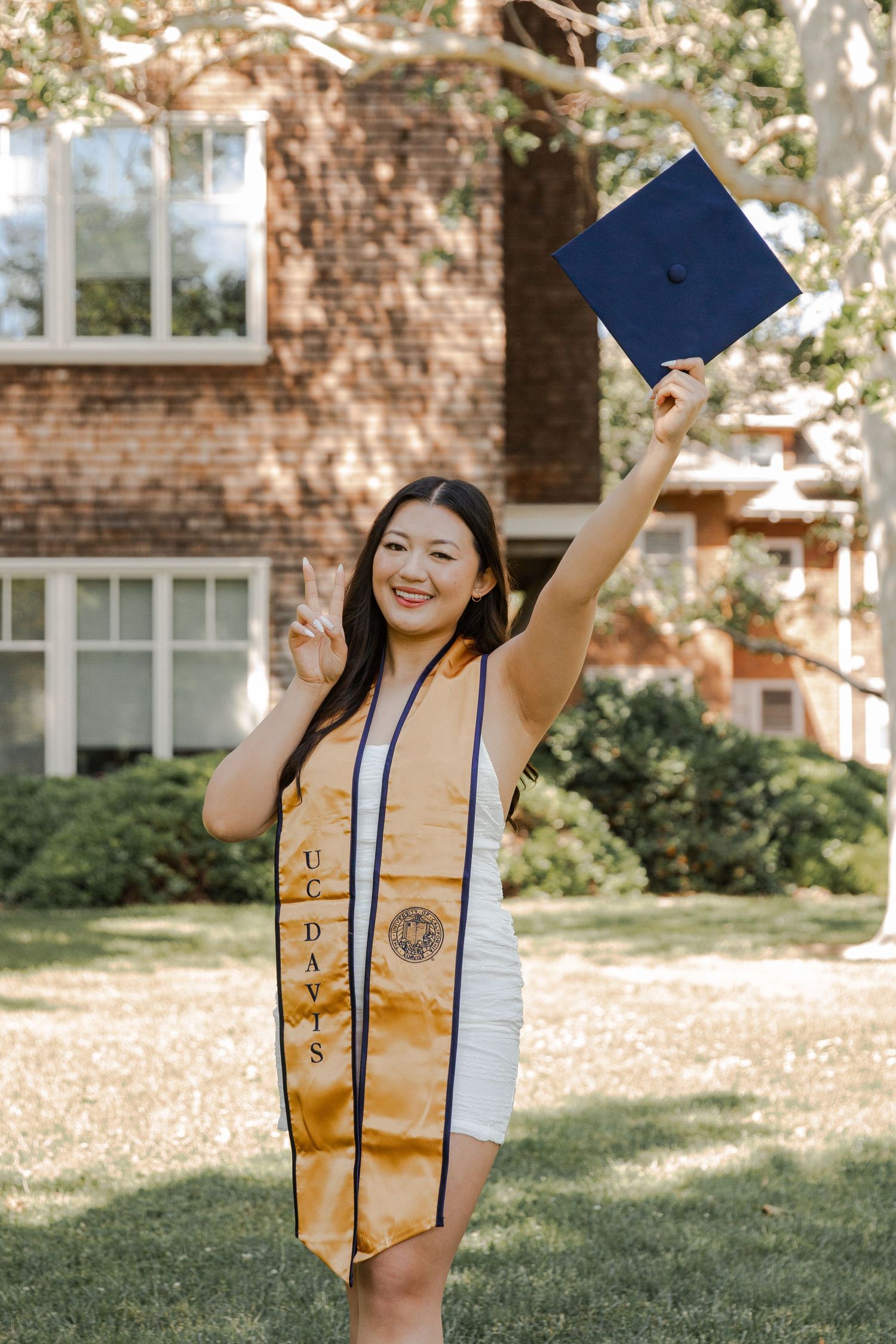 Graduate in white dress holds blue cap celebrating on campus lawn.