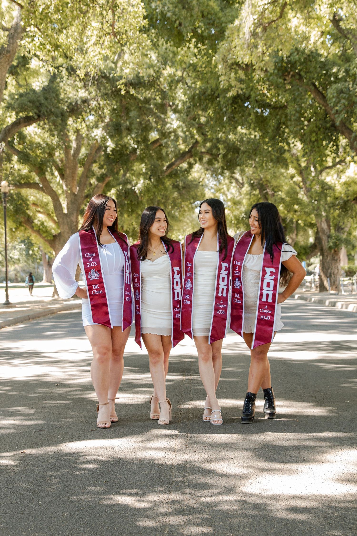 Four graduates in white dresses and red sashes pose together on a tree-lined path.