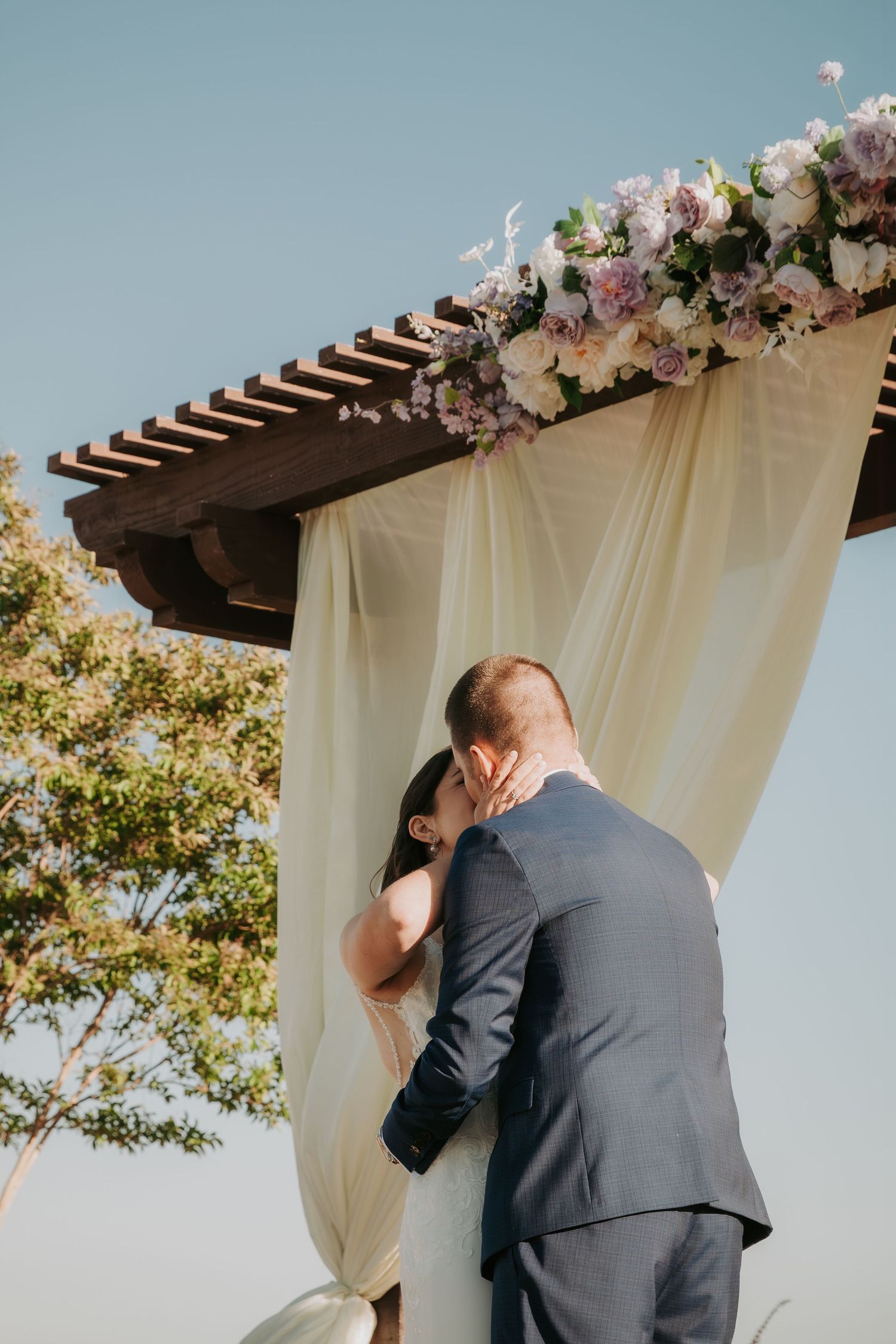 Romantic wedding ceremony under a wooden pergola decorated with white drapes and pink floral arrangements.