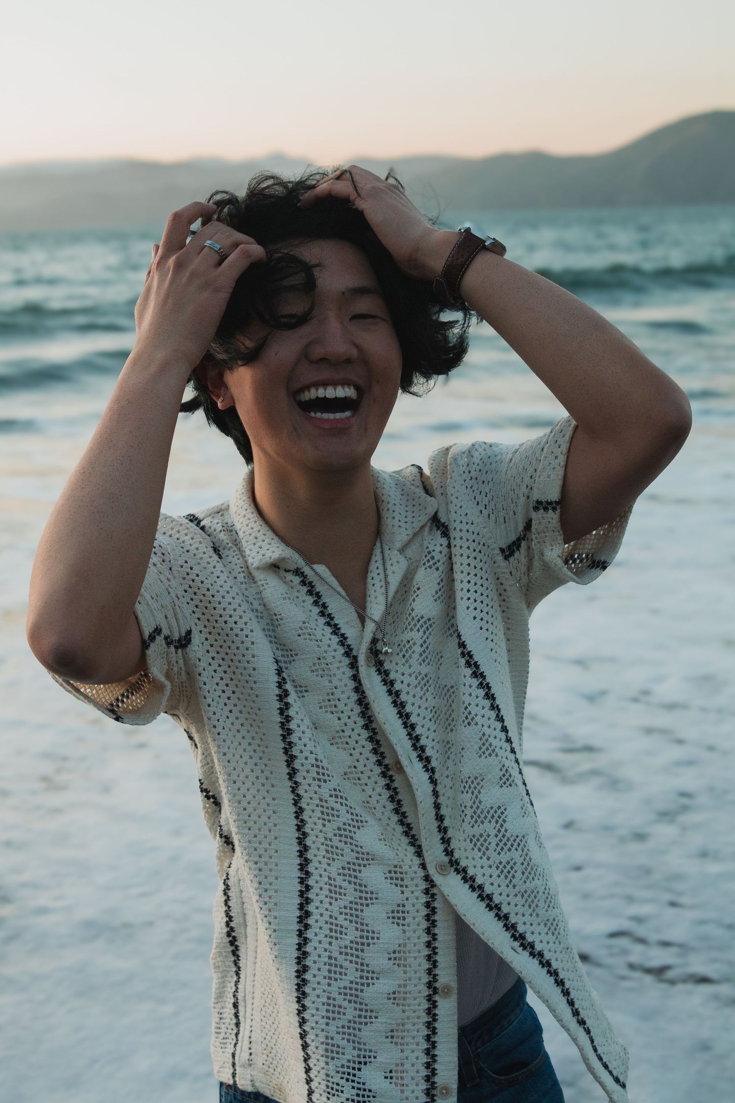 Person laughing joyfully at sunset on a beach wearing a patterned white shirt with ocean waves in the background.