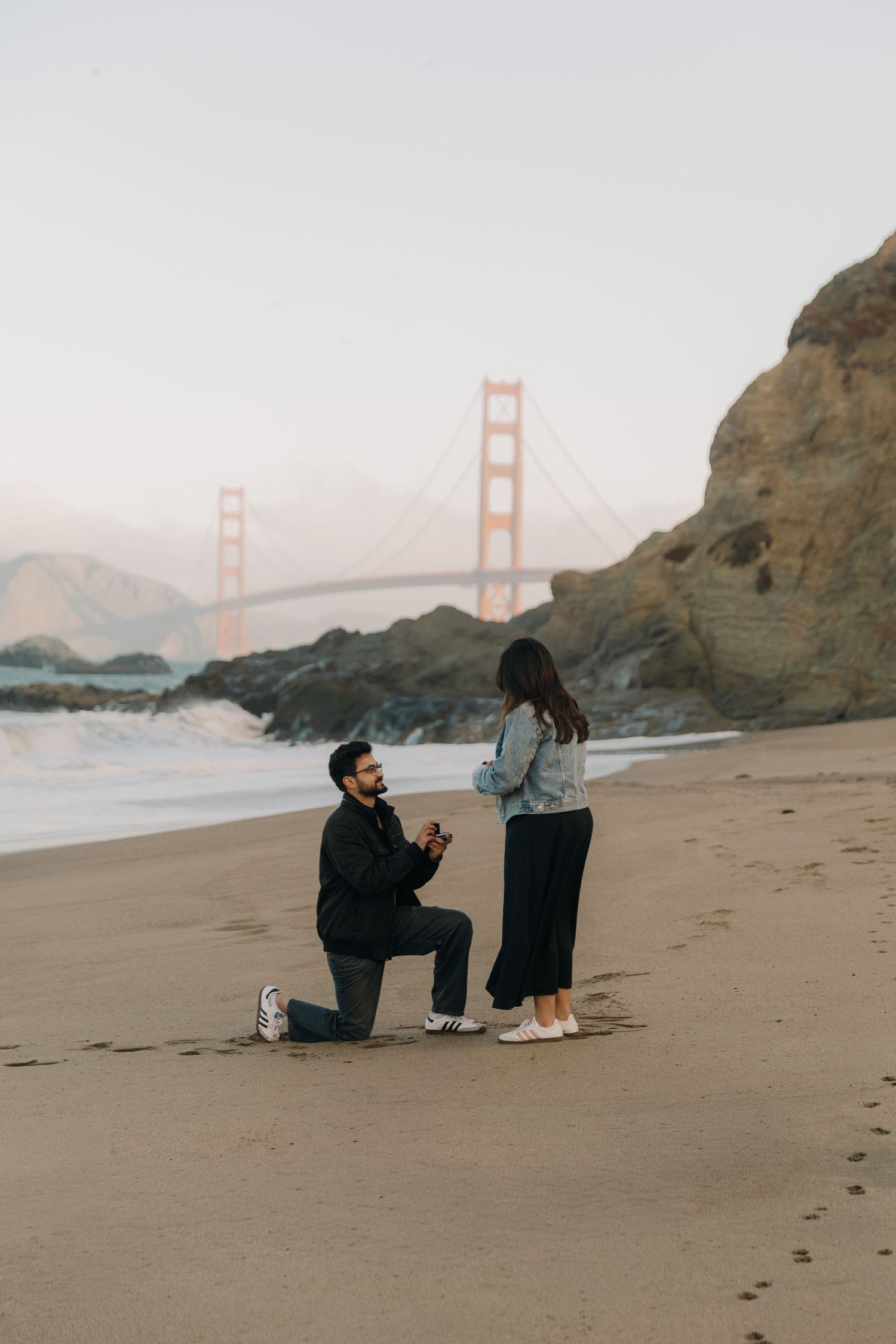 A couple shares a romantic moment on Baker Beach with the Golden Gate Bridge visible in the background at sunset.