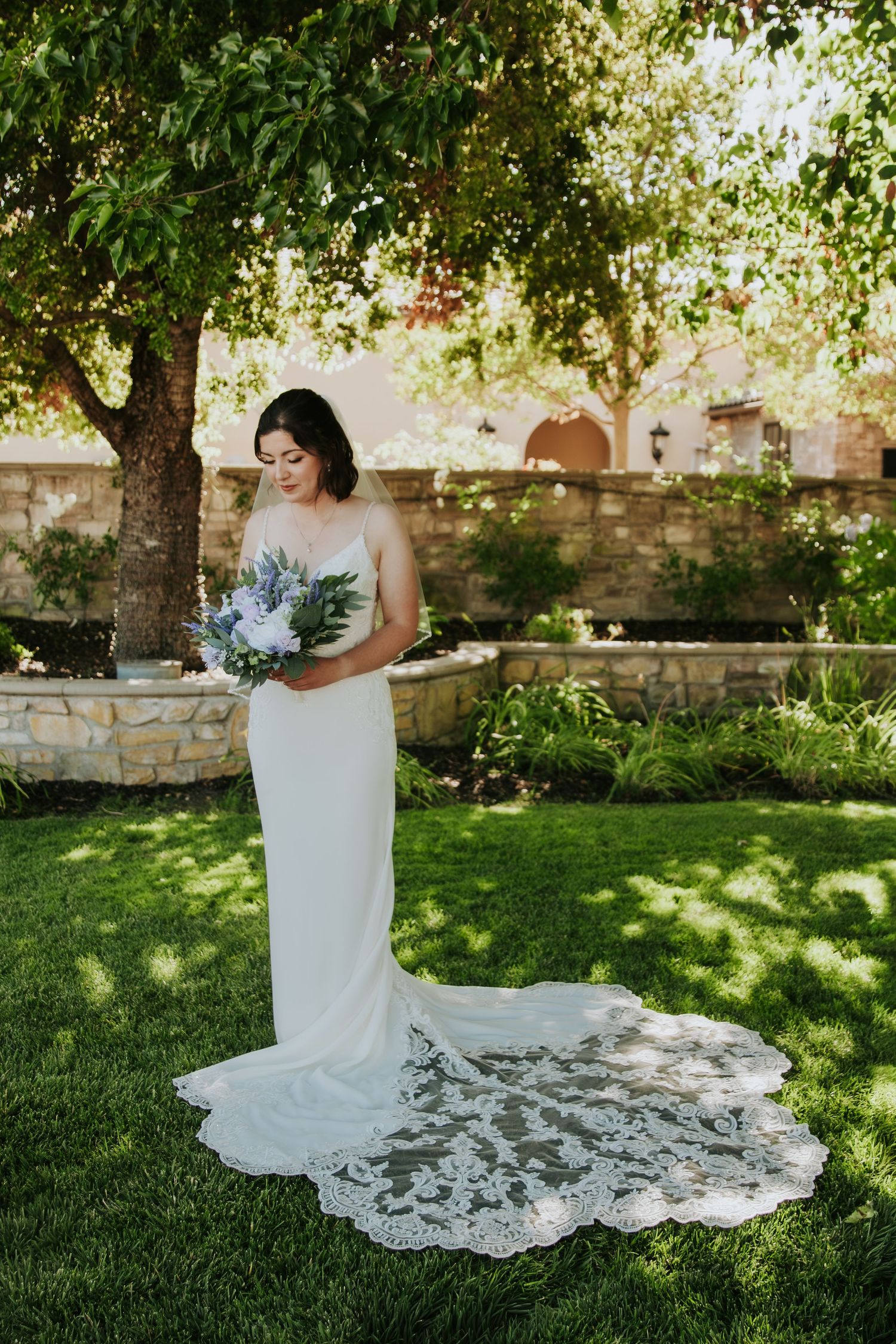 A white wedding dress with a long lace train flows across green grass in a garden setting with stone walls and trees.