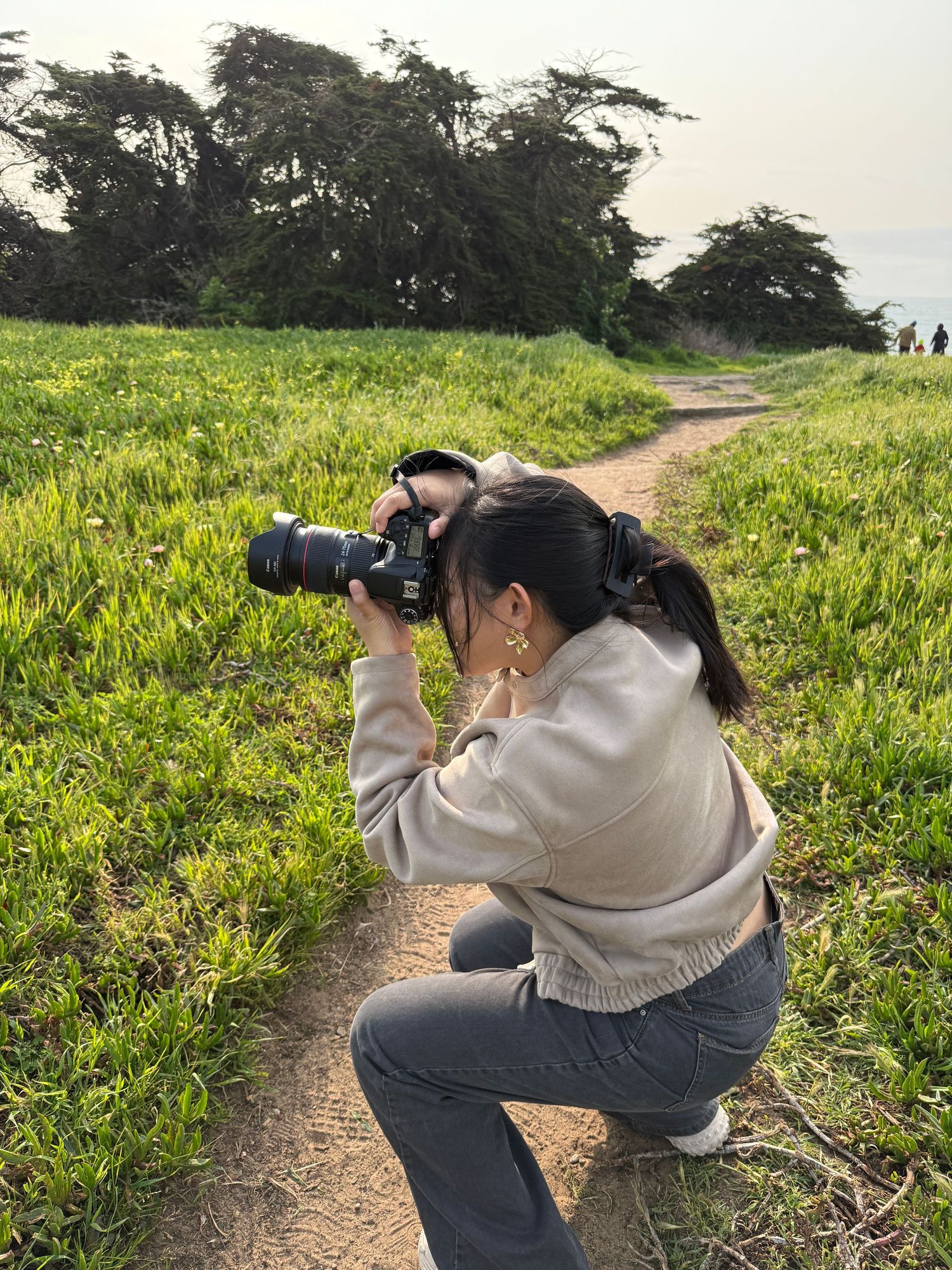 A behind-the-scenes photo of Allyson crouching on a dirt path amid yellow wildflowers taking photos in Thorton Beach.