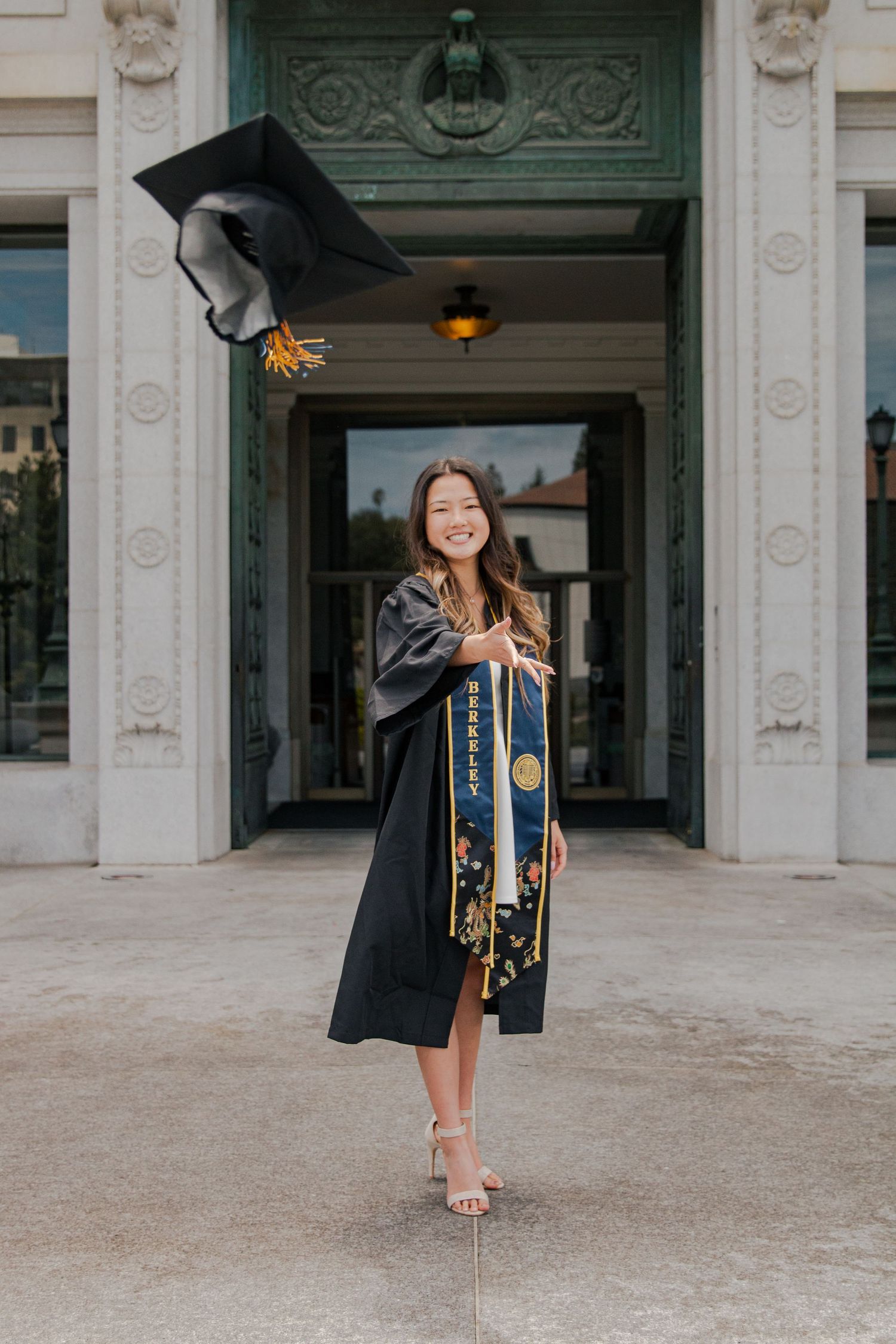 Graduate in black cap and gown poses elegantly in front of ornate building entrance.