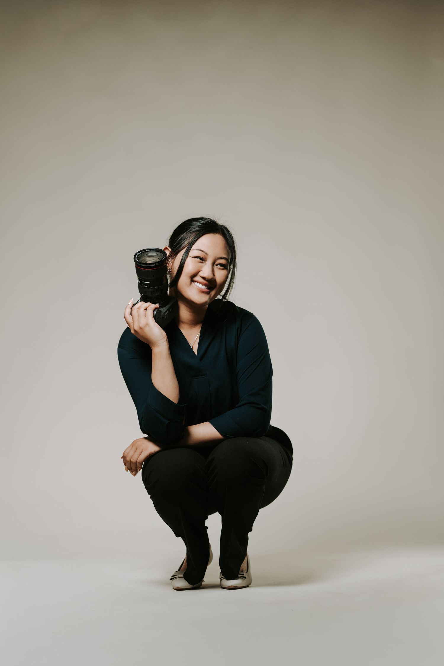 A photographer in all black crouches in various poses against a light neutral backdrop while holding a camera.