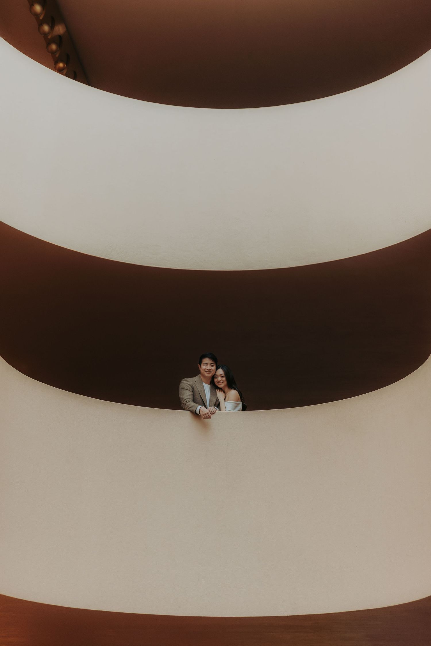 A couple embraces in a modern architectural space with curved white and brown walls creating dramatic lines and shadows.