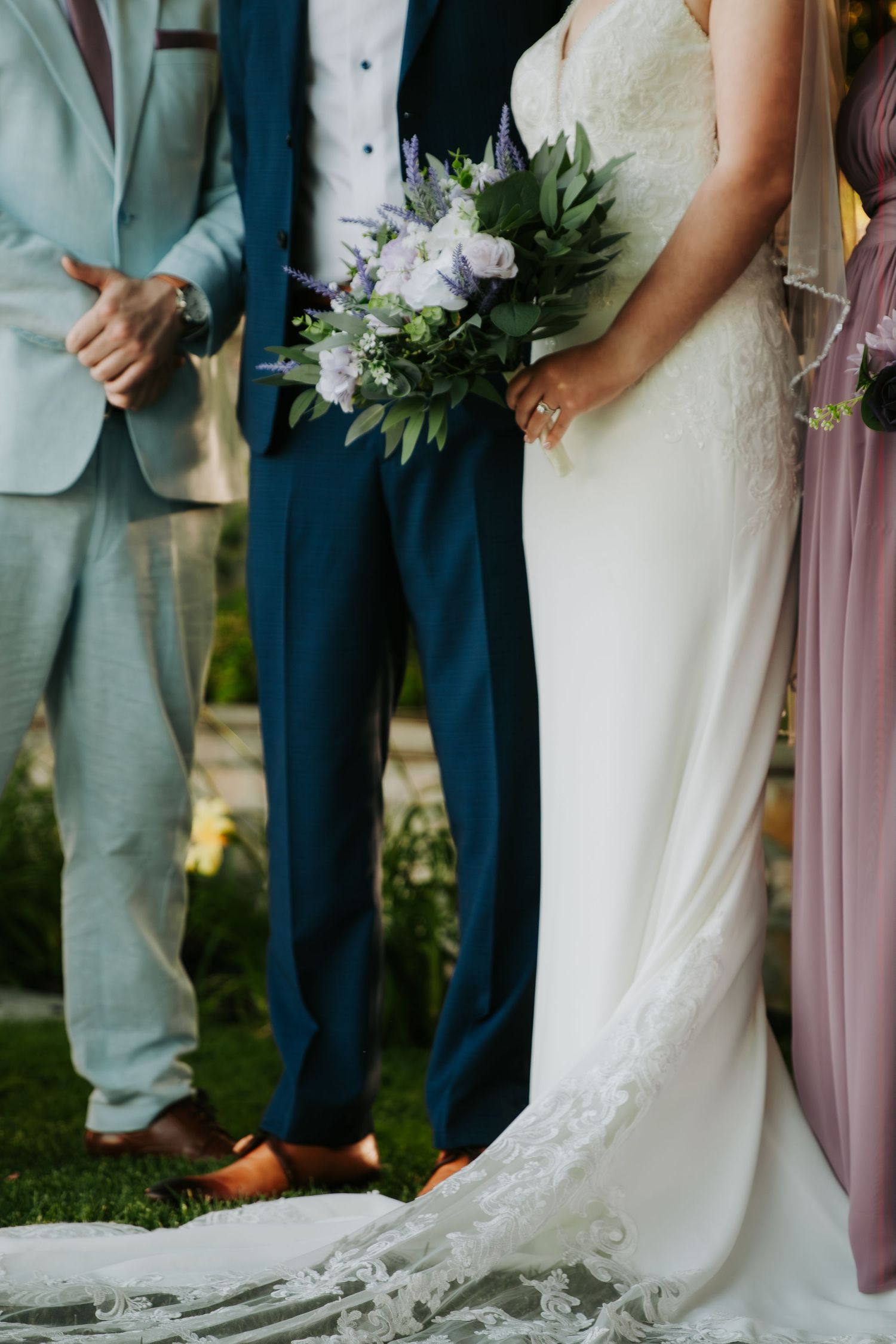 Wedding party stands in a line showing elegant white bridal gown train details and groomsmen in navy suits.