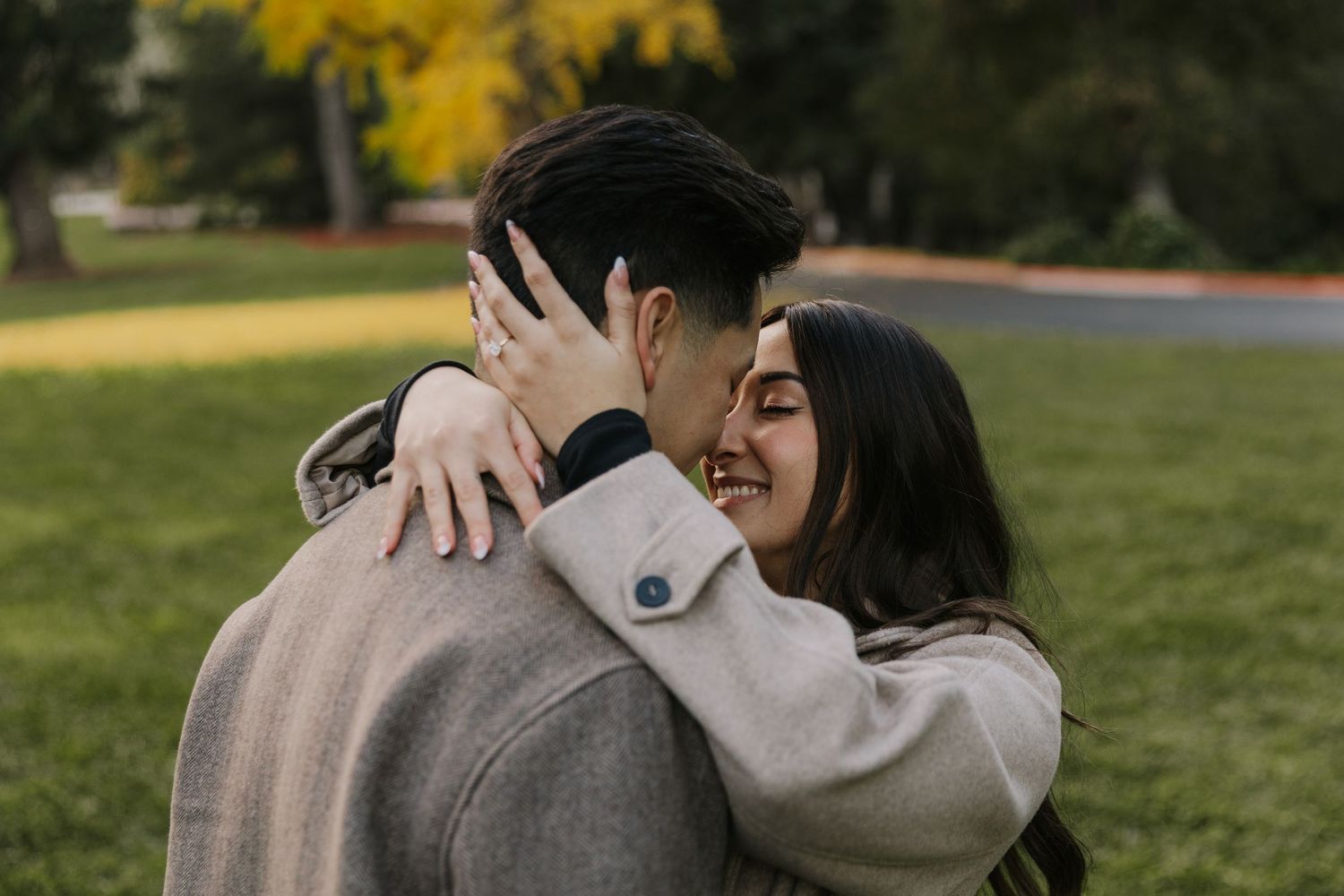 A couple embraces tenderly in a park with autumn foliage in the background.