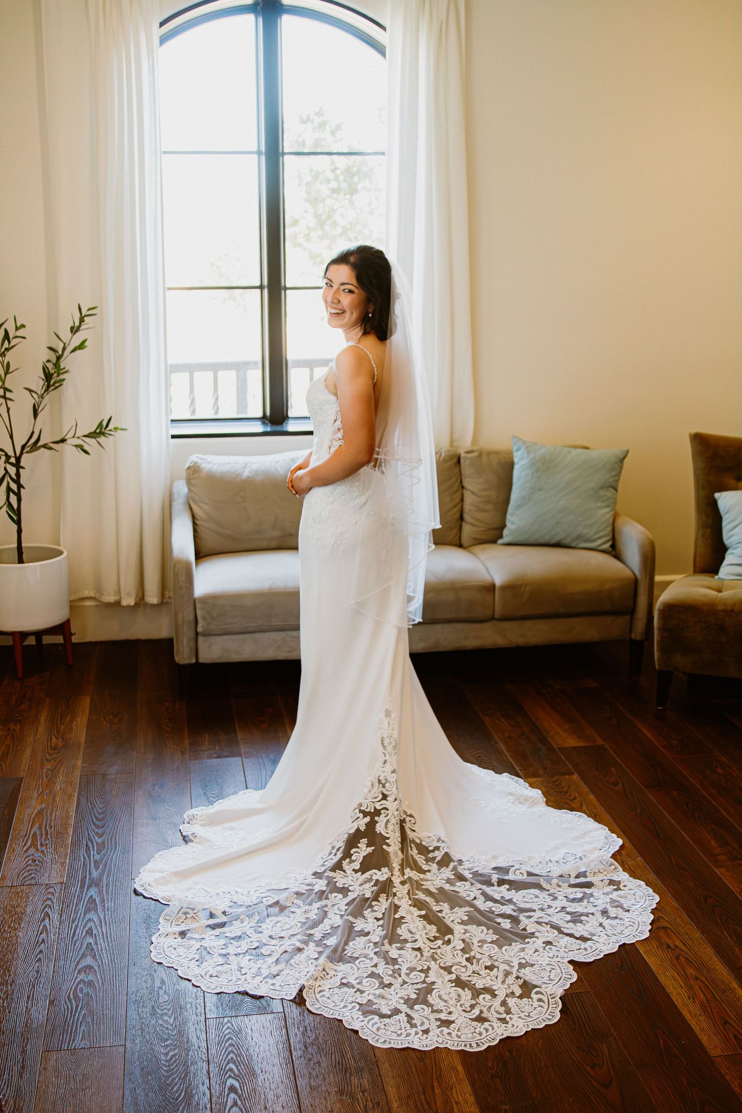 A wedding dress with intricate lace train displayed on hardwood floors in natural window light.