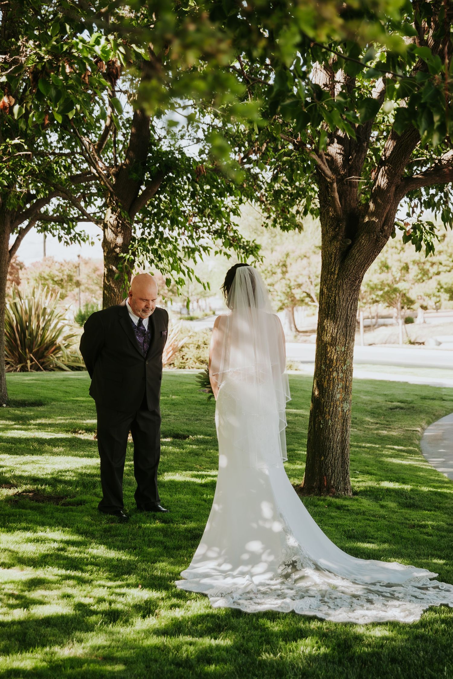 A romantic first look moment between newlyweds in a sun-dappled garden setting with green trees and grass.