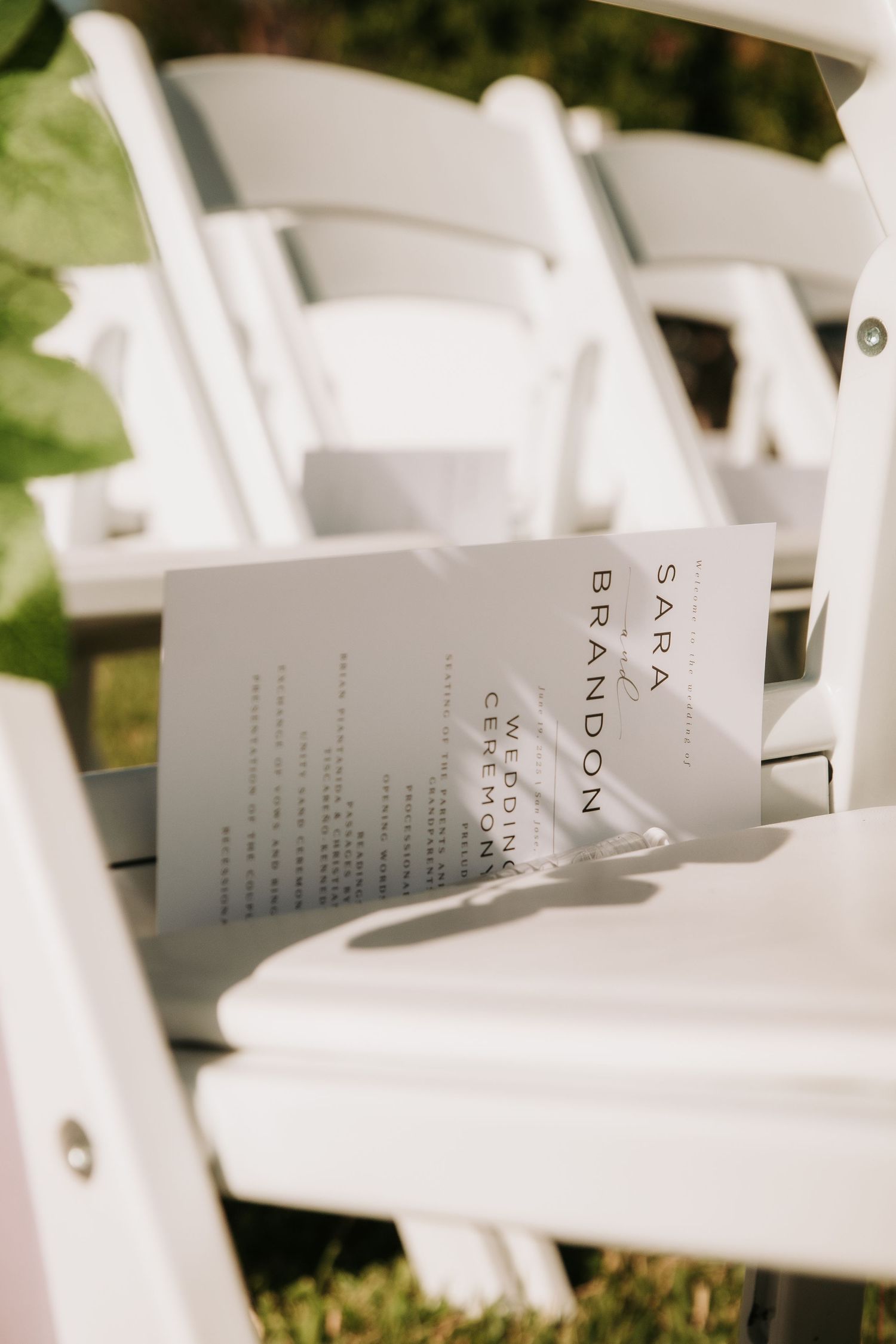 A wedding ceremony program rests on a white folding chair outdoors.
