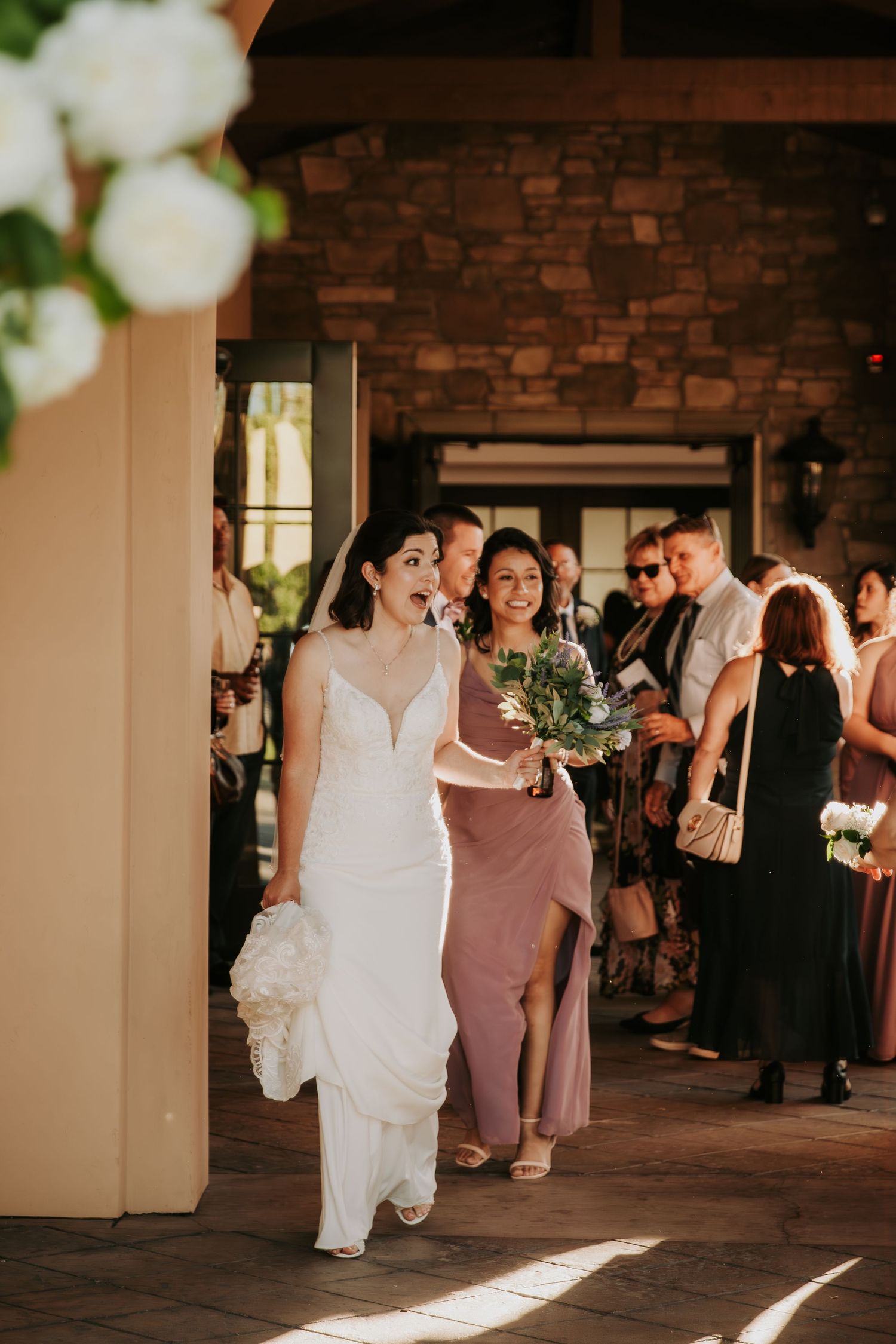 Wedding party dances and celebrates in a rustic brick-walled reception venue with natural light streaming in.