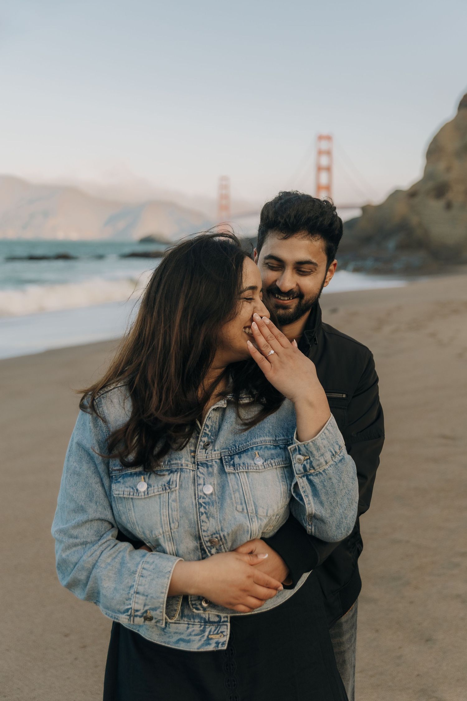 A couple shares intimate moments at a beach with the Golden Gate Bridge visible in the background at sunset.