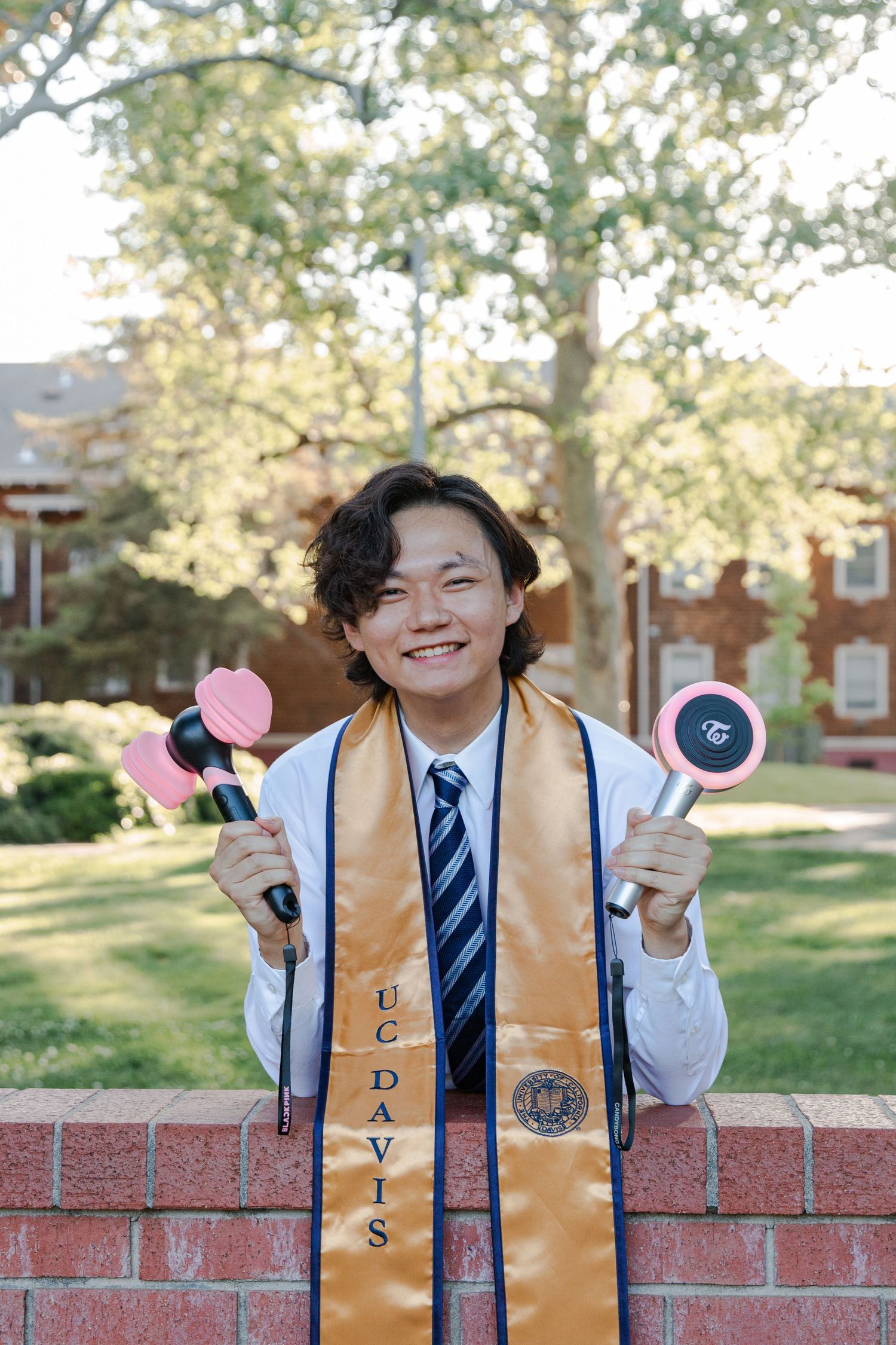 Graduate in formal attire holds decorative props while posing with gold stole on brick wall.