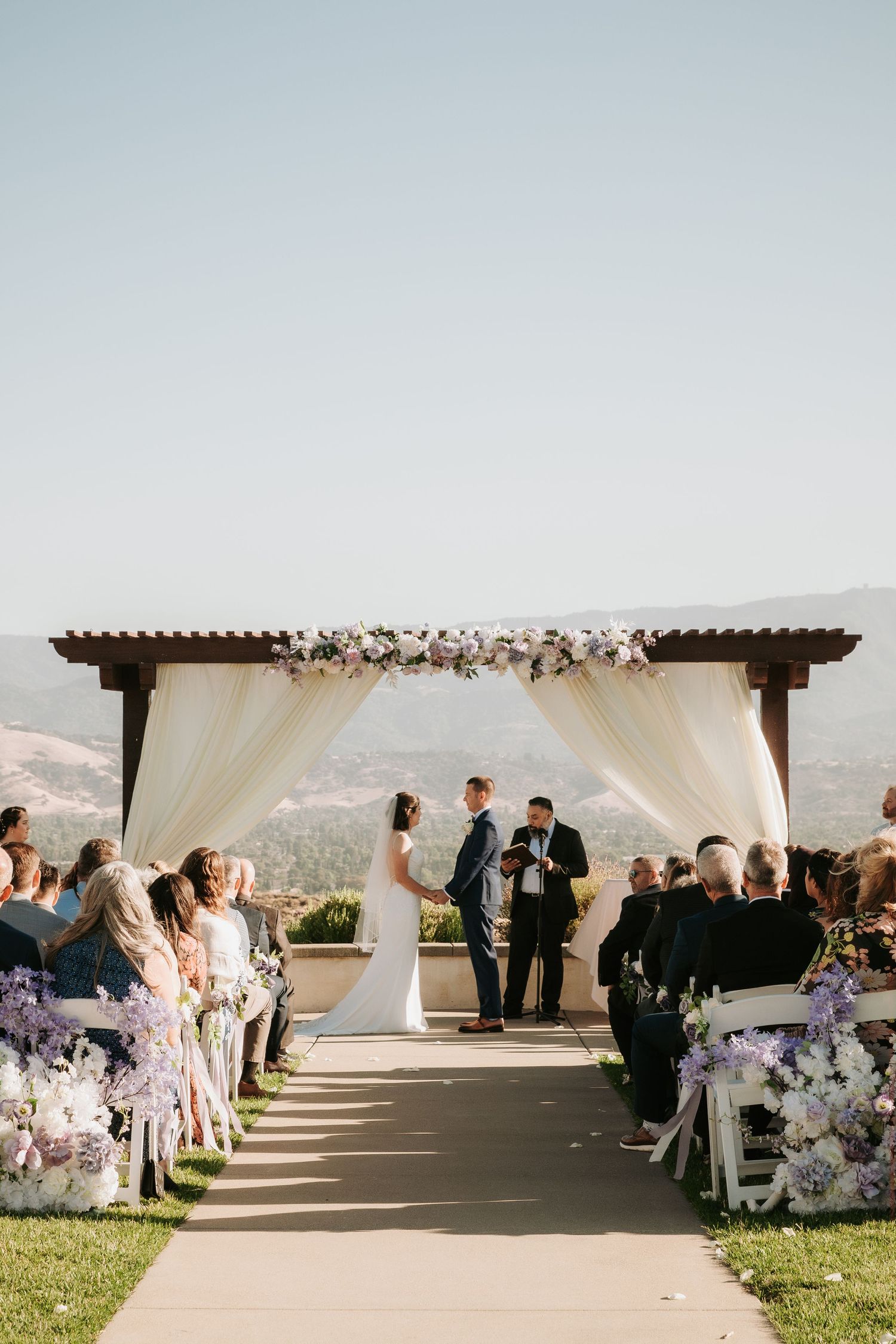 An outdoor wedding ceremony on a hilltop with white draping, floral arrangements and guests seated on either side of the aisle.
