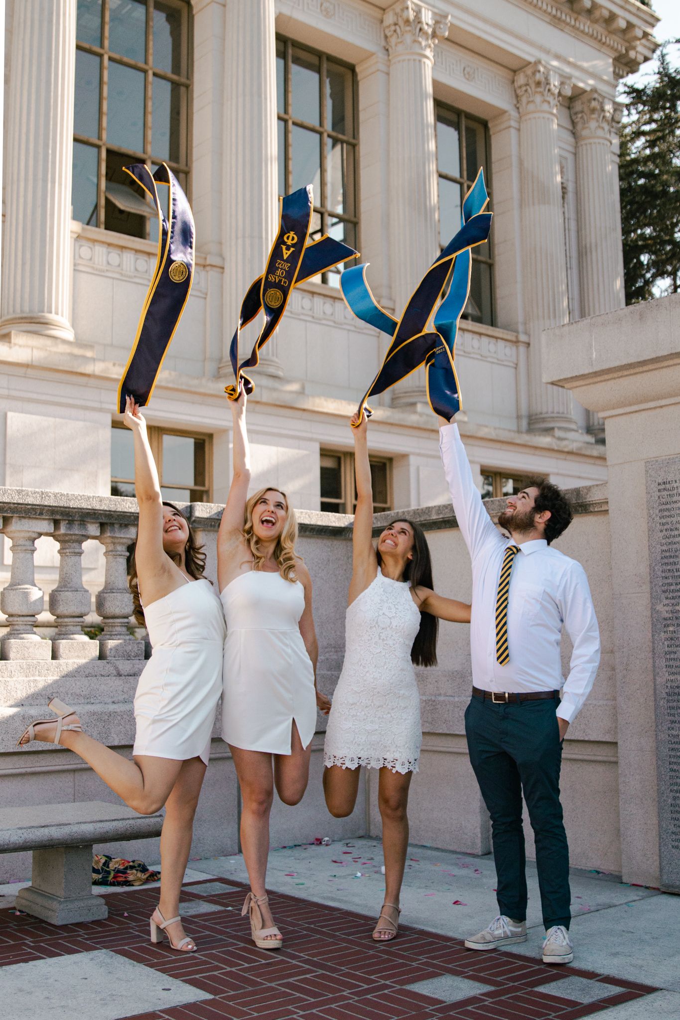 Three graduates throw their sashes in the air while celebrating on campus steps.