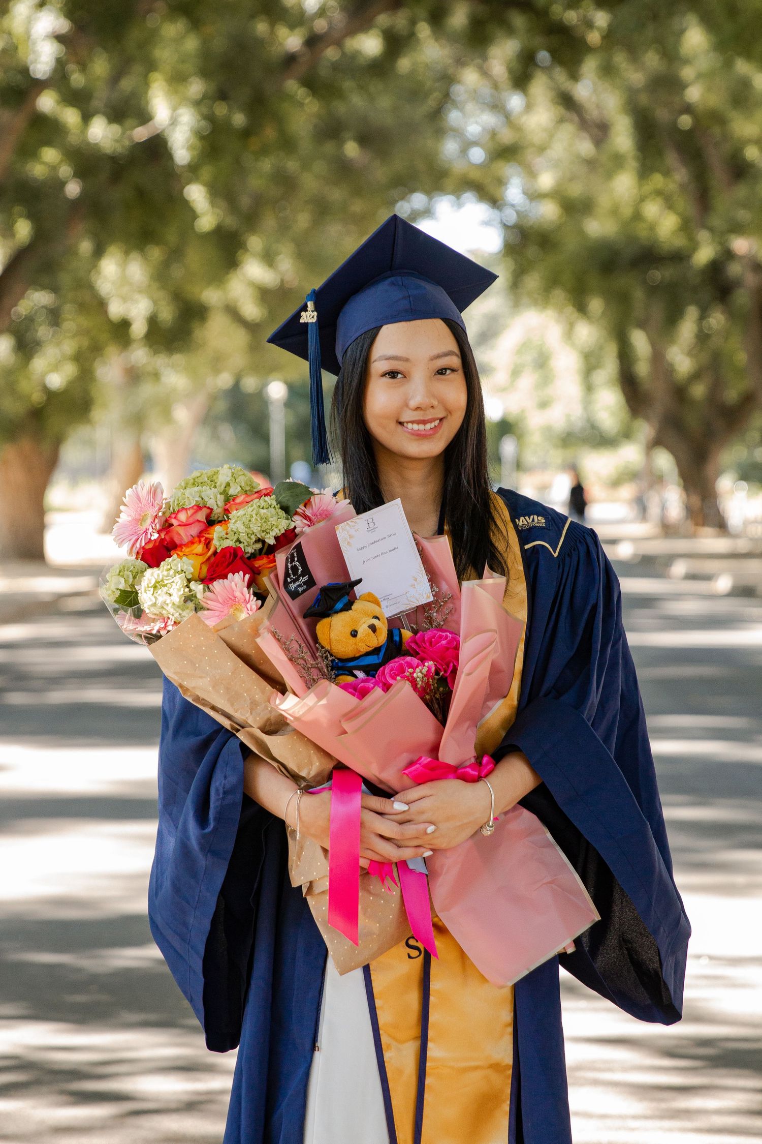 Graduate in navy cap and gown holds colorful bouquets during outdoor celebration.