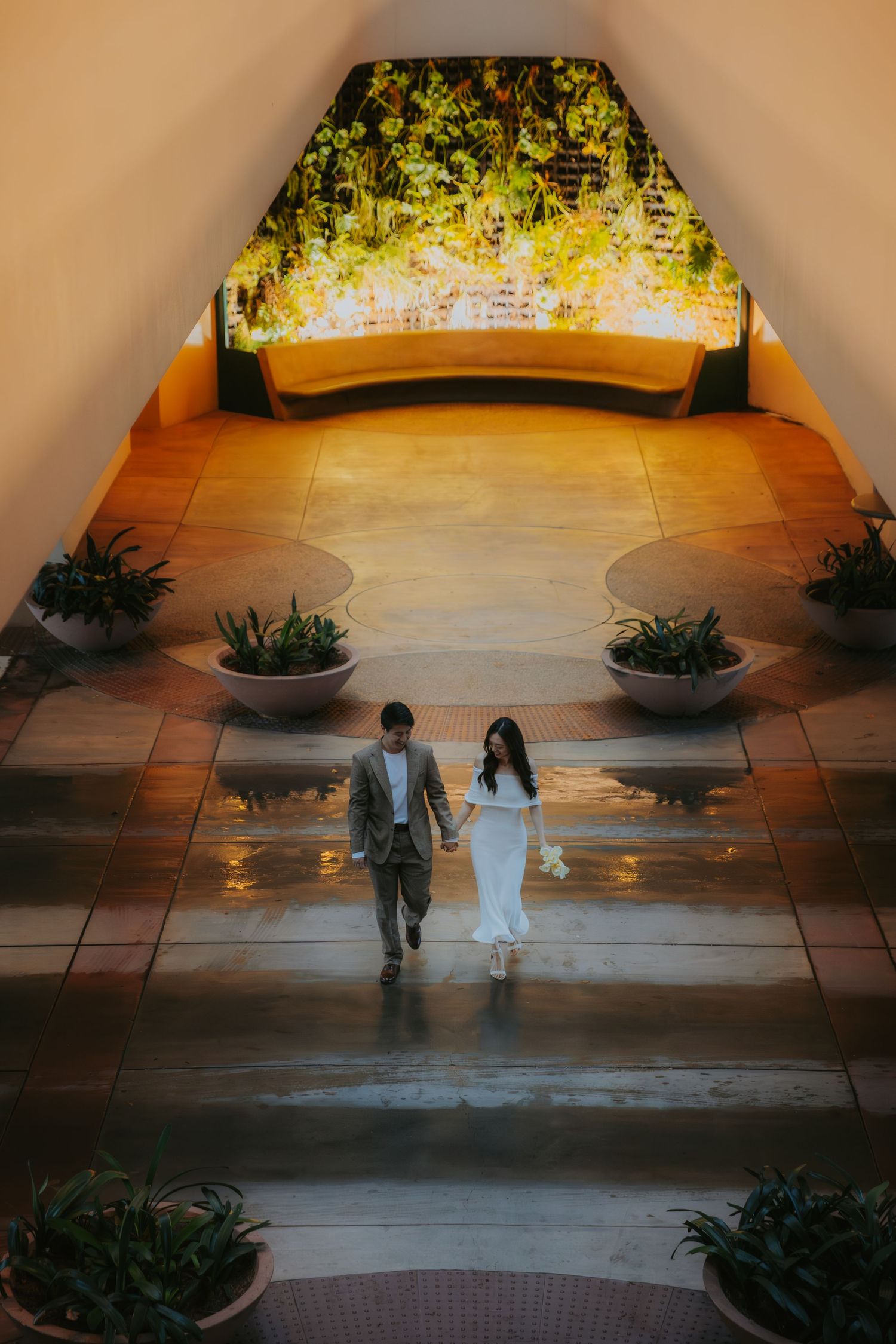 Two figures cast long shadows on a tiled patio at sunset, with potted plants framing the edges of the scene.