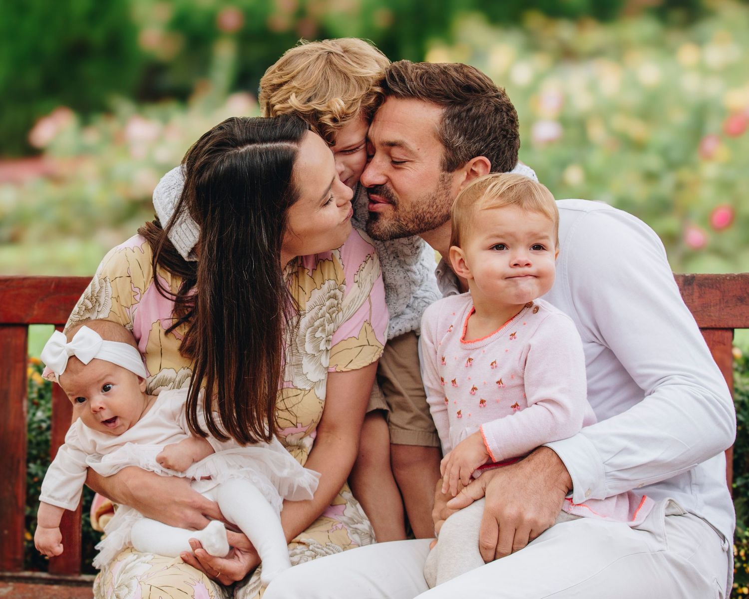 A family shares an intimate moment together while sitting on a wooden bench in a garden with blooming flowers.