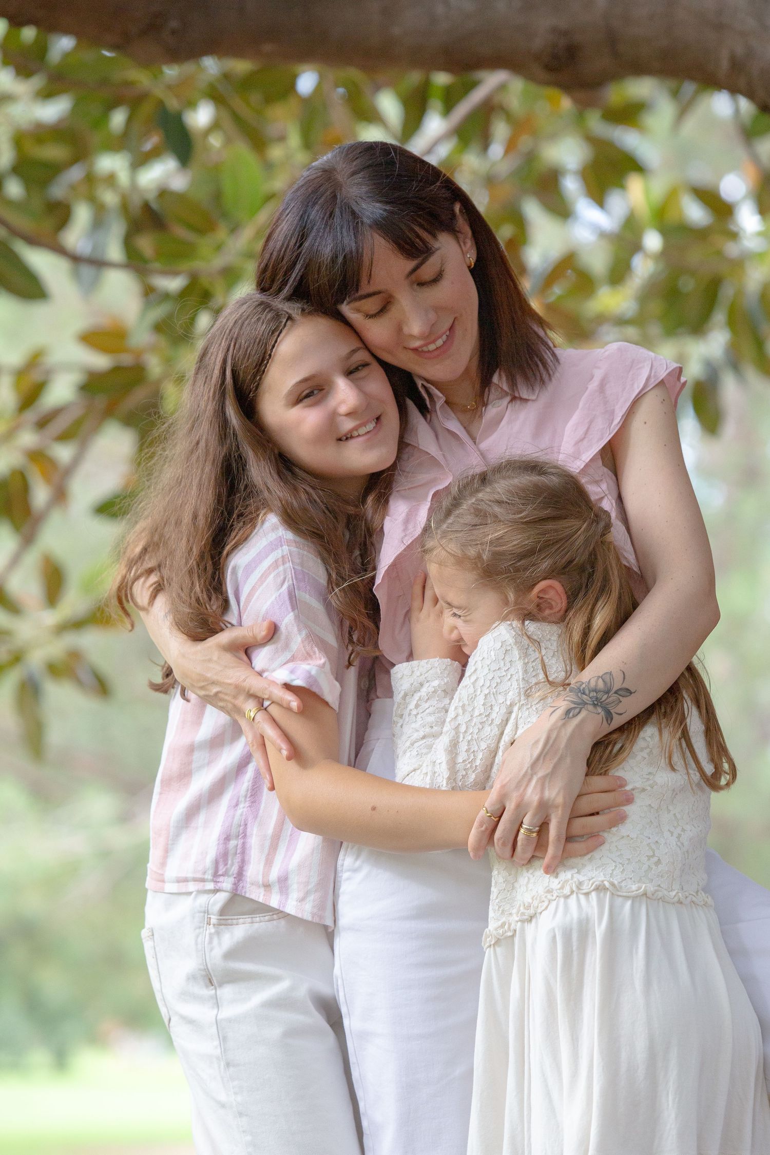A mother tenderly embraces her two daughters outdoors under a tree, all wearing white and pink outfits.