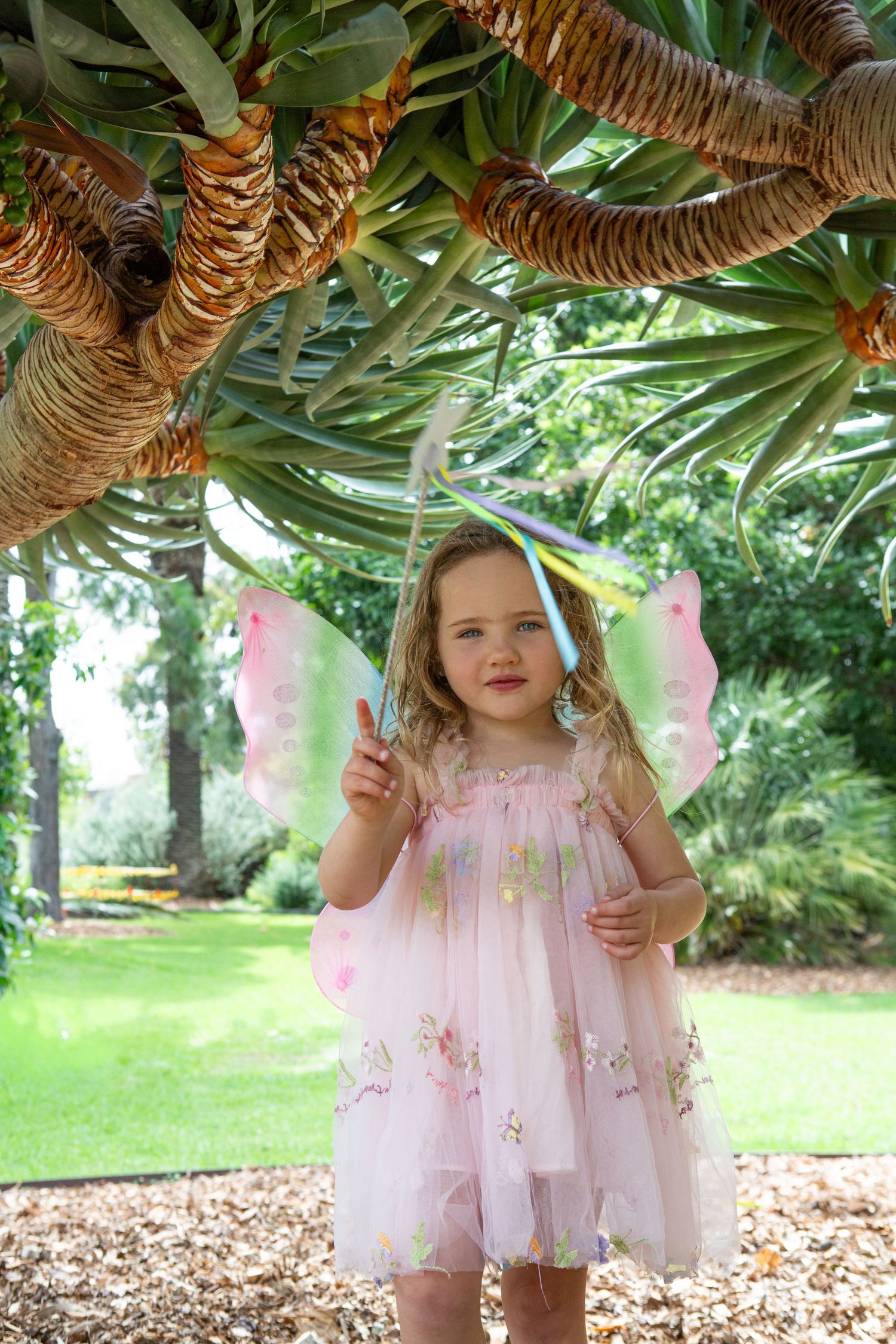 A little girl in a pink fairy costume with butterfly wings holds a wand under a tropical tree in a lush garden.