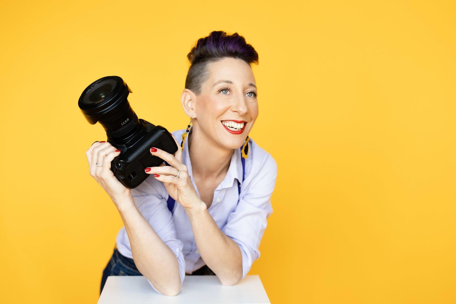 Professional photographer holding DSLR camera and smiling against bright yellow background.