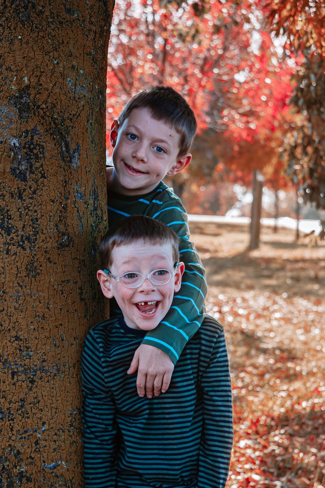 Two children pose playfully by tree trunk with autumn foliage in background.