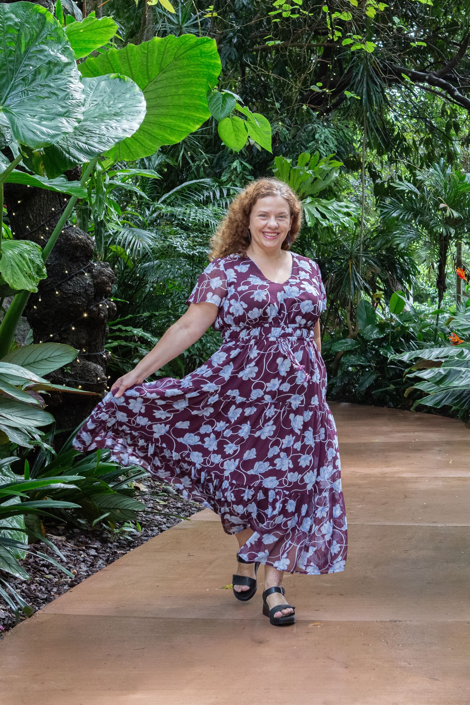 Woman in floral maxi dress walking through lush tropical garden path, smiling and holding her skirt.