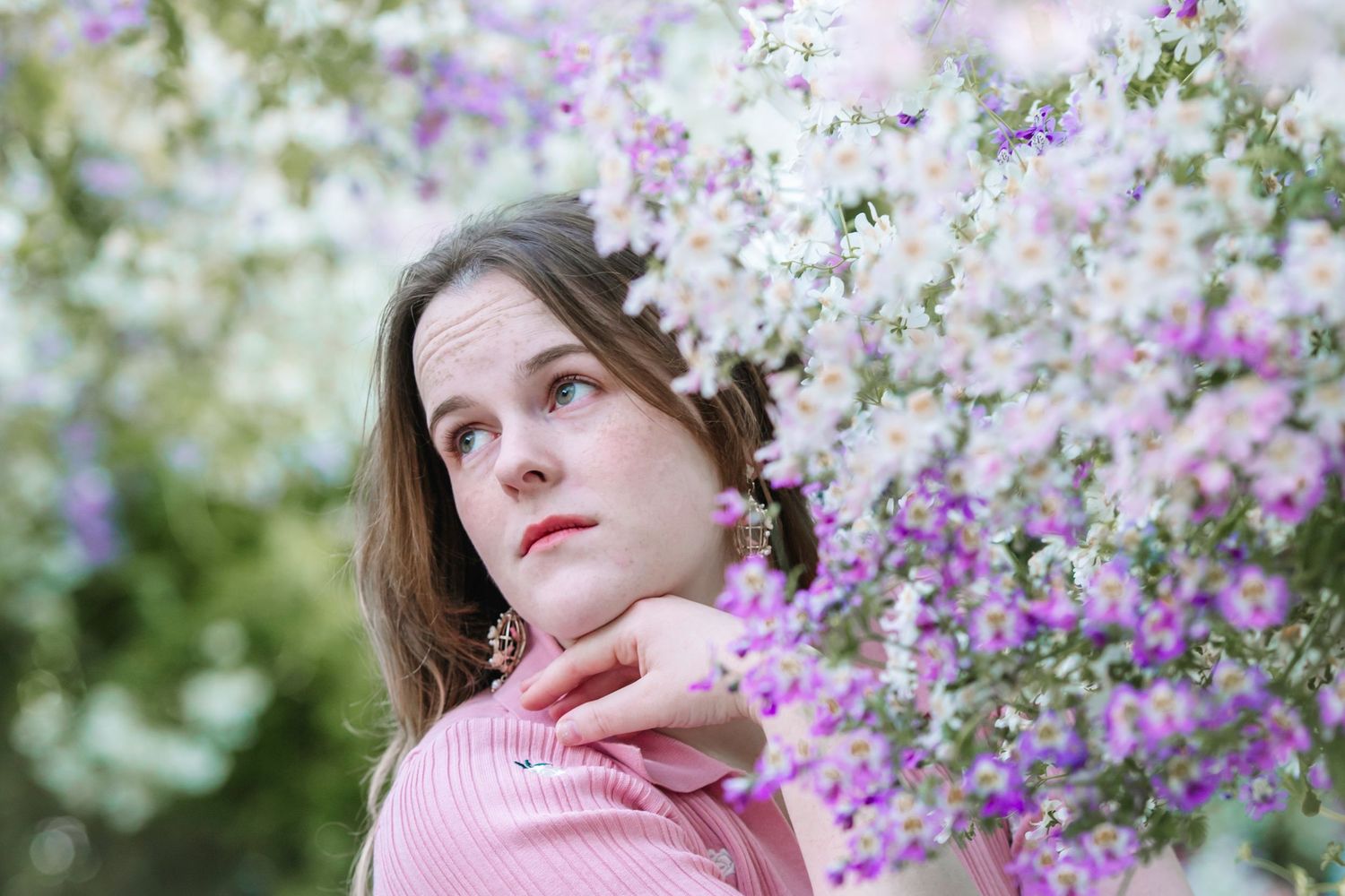Young woman in pink top poses thoughtfully among blooming purple and white flowers in a garden setting.