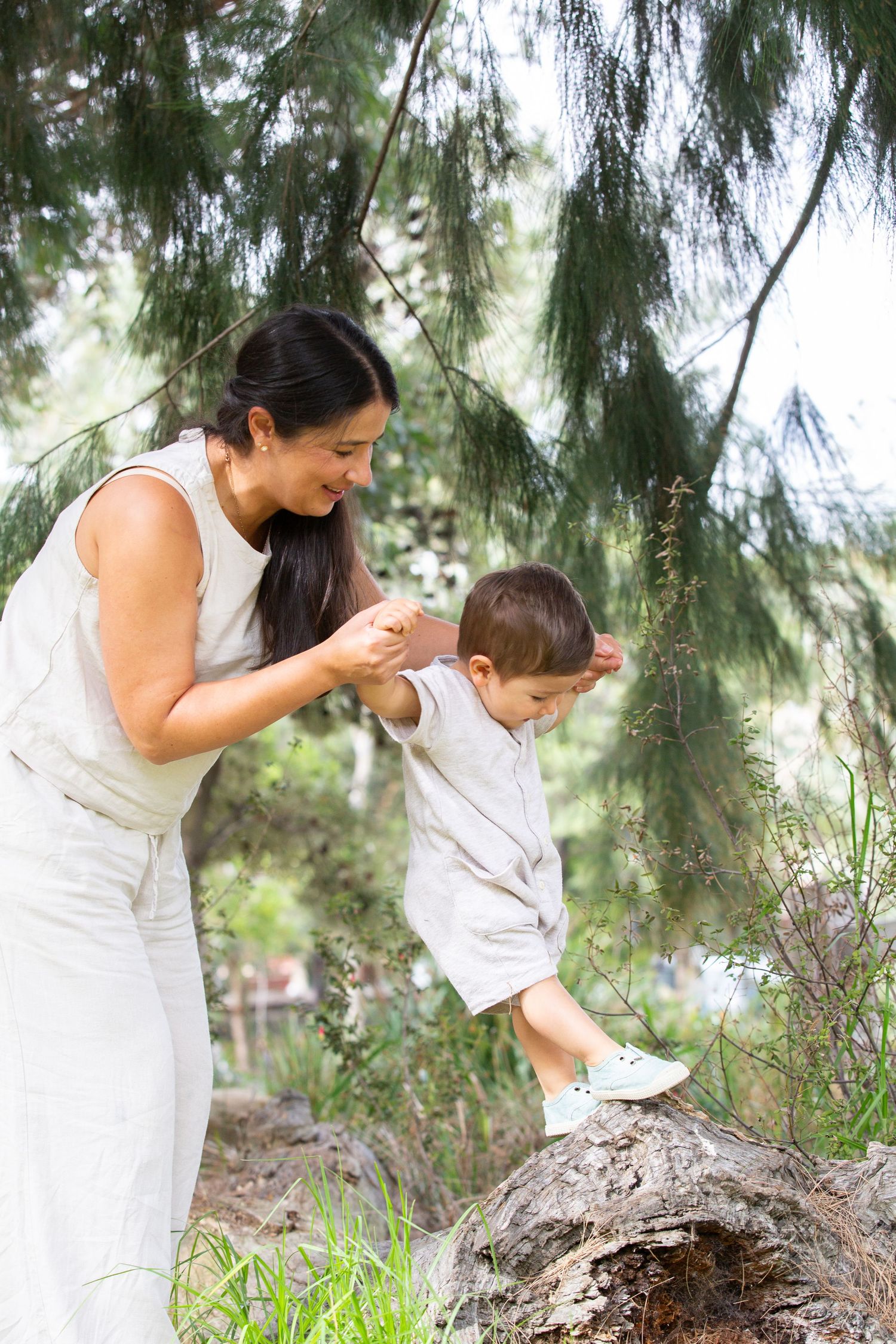 A smiling mother helps her toddler climb a log in a lush green park, both wearing matching neutral outfits.