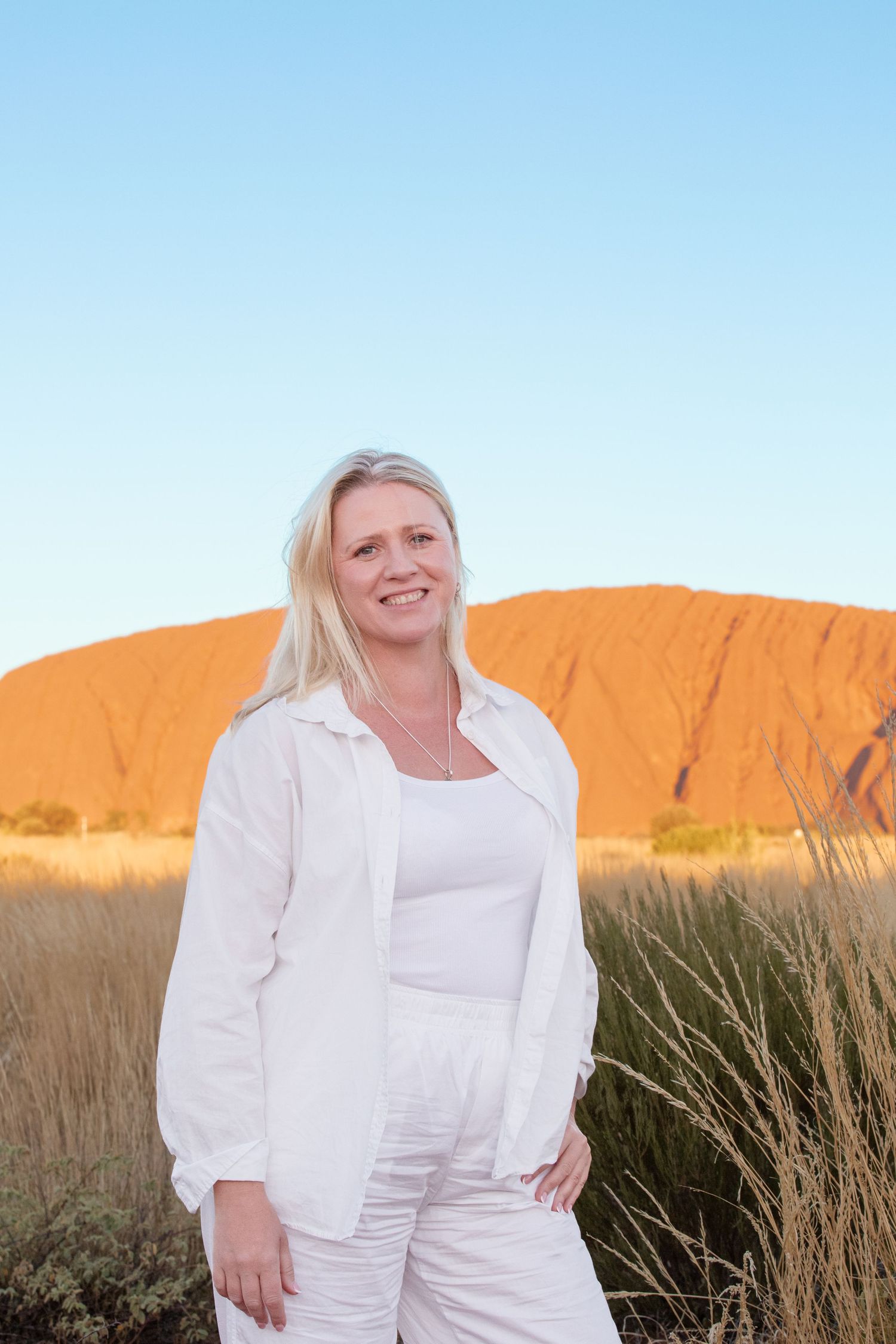 Tourist in white outfit poses in front of Uluru rock formation during sunset in Australia's Northern Territory.