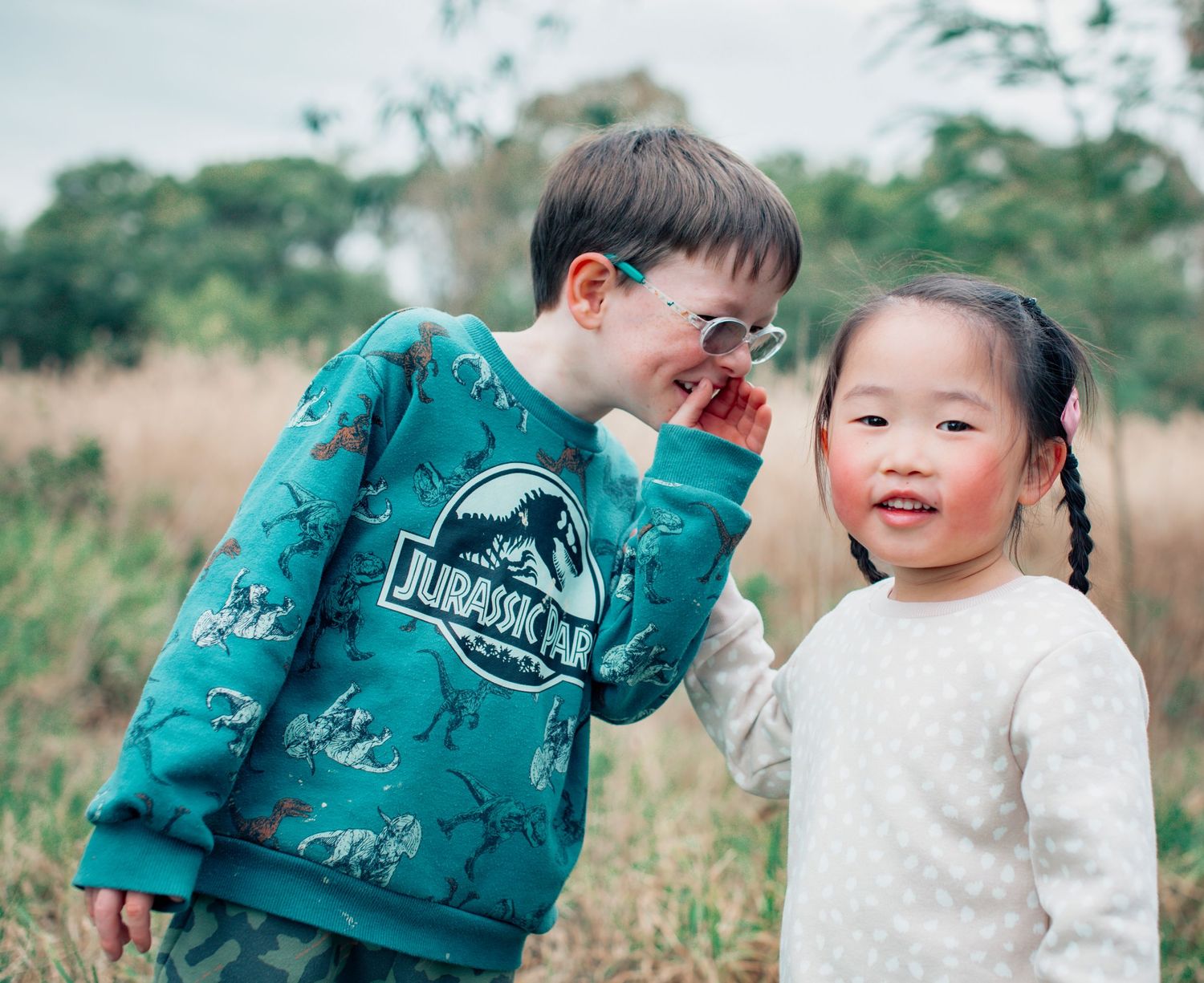 Two children sharing a laugh together outdoors, one wearing a dinosaur-themed sweatshirt.