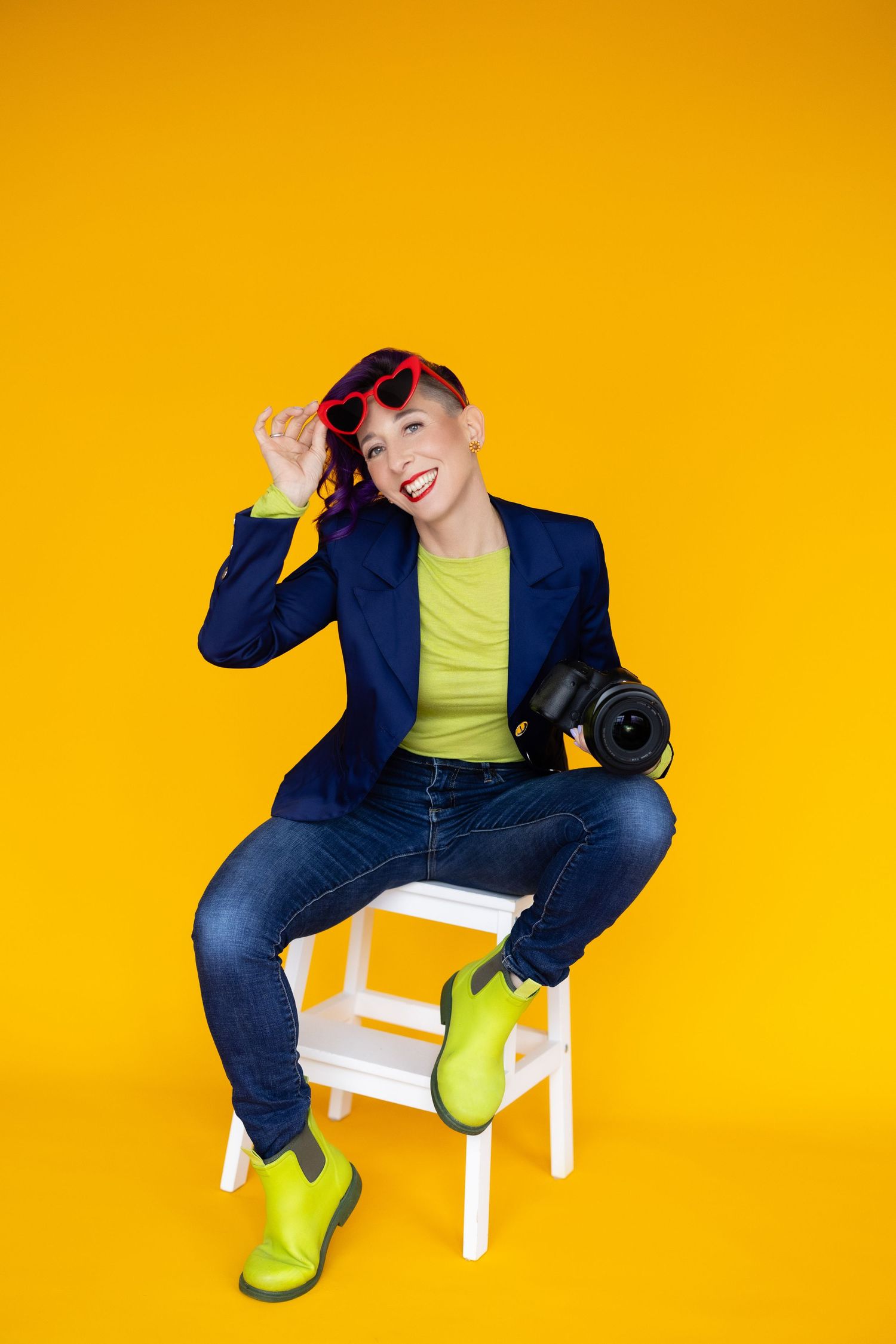 Woman sitting on white stool holding camera, wearing blue blazer, green boots, and red heart sunglasses on yellow background.