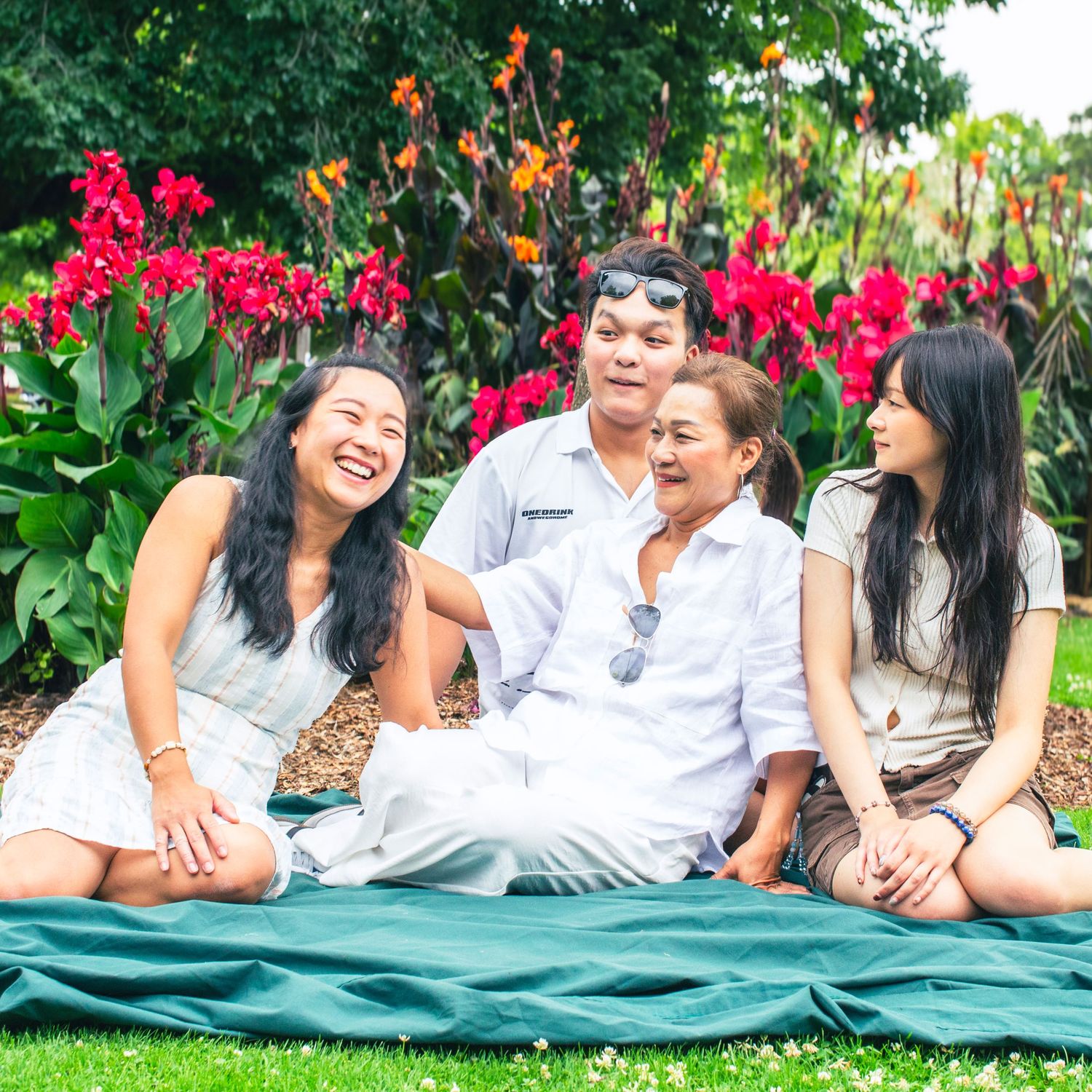 Family of four sitting on green blanket laughing together surrounded by vibrant tropical flowers in a garden.