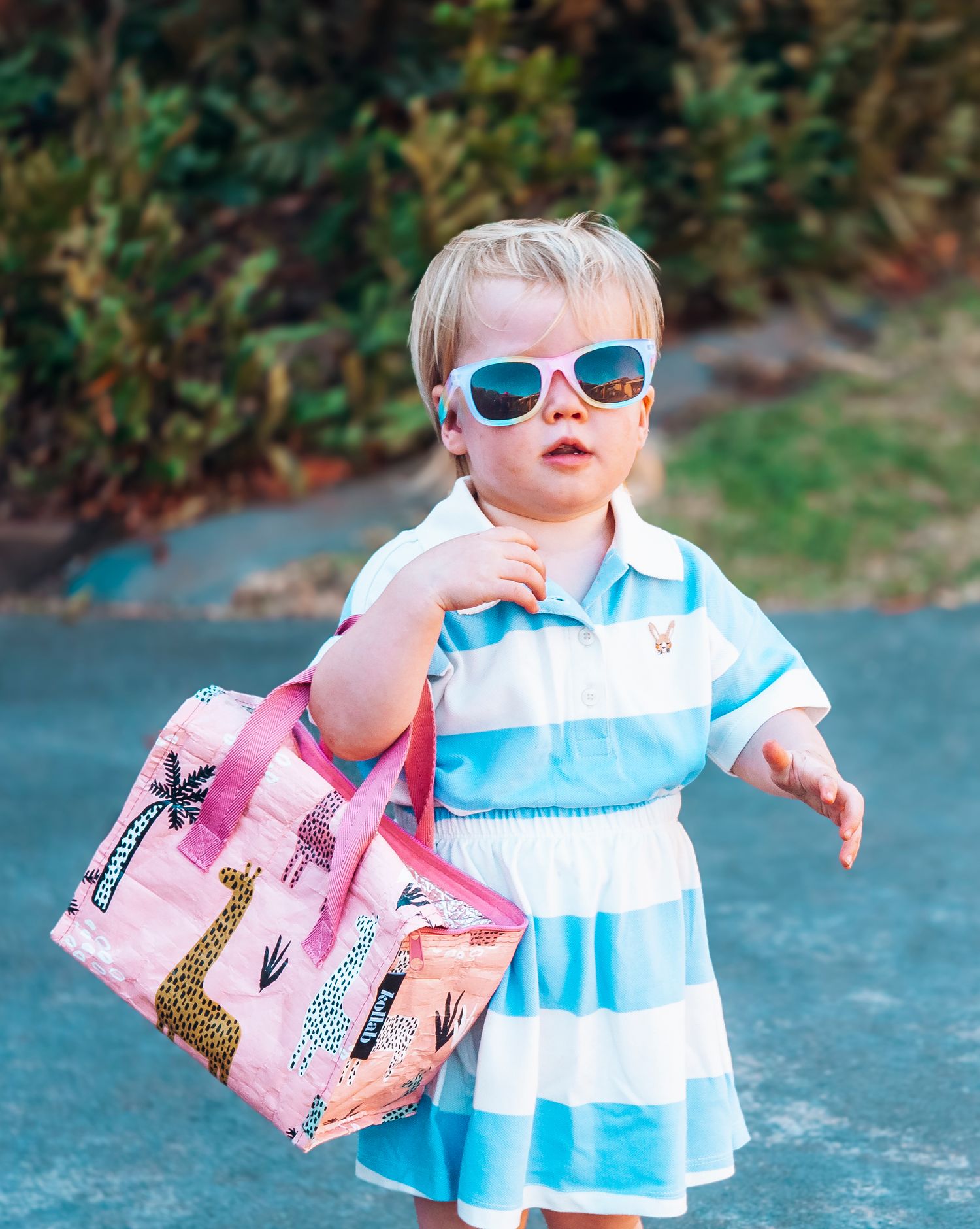 Child in striped dress and sunglasses carrying pink shopping bag outdoors.