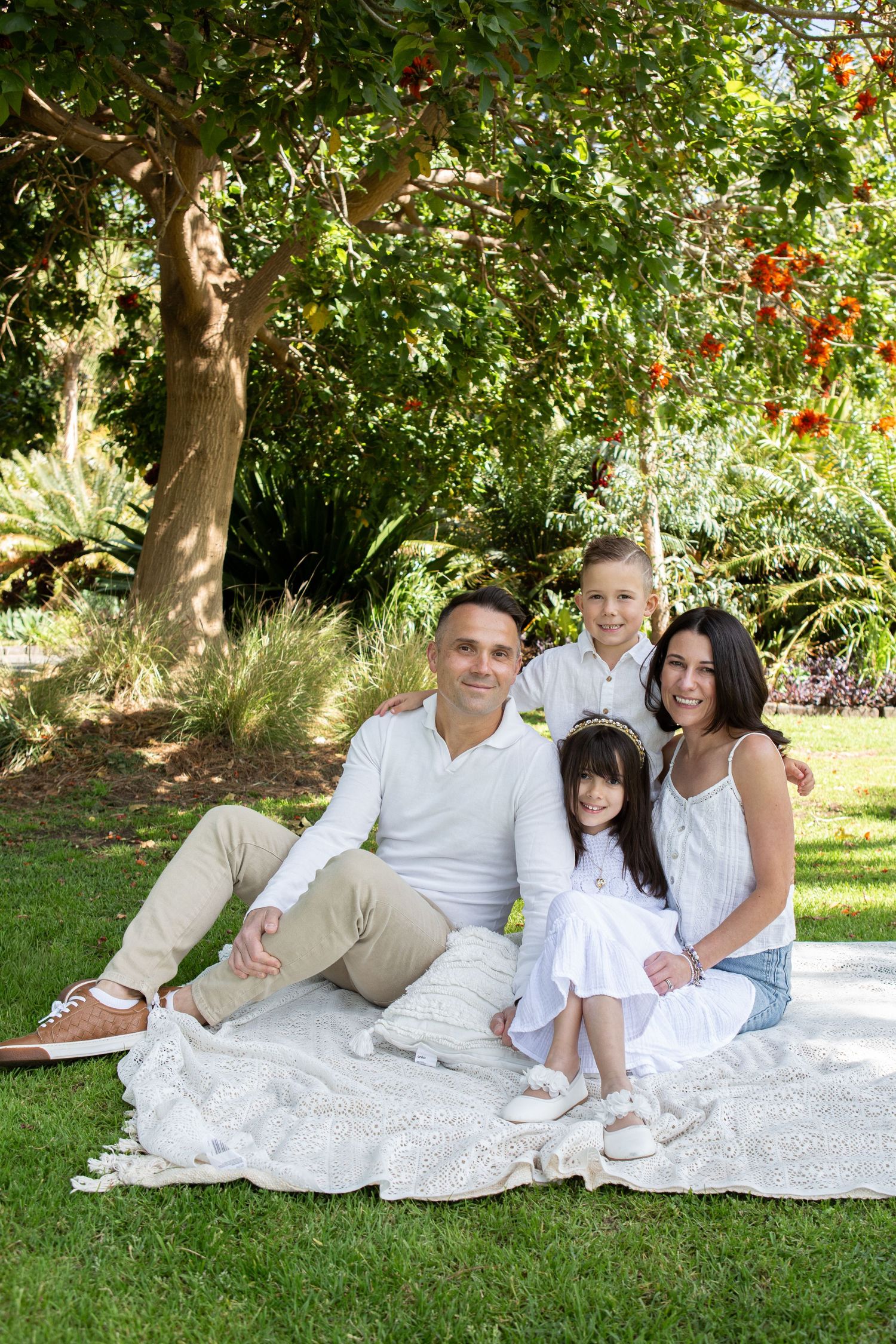 A family wearing white and beige clothing relaxes together on a white blanket in a sunny garden setting.