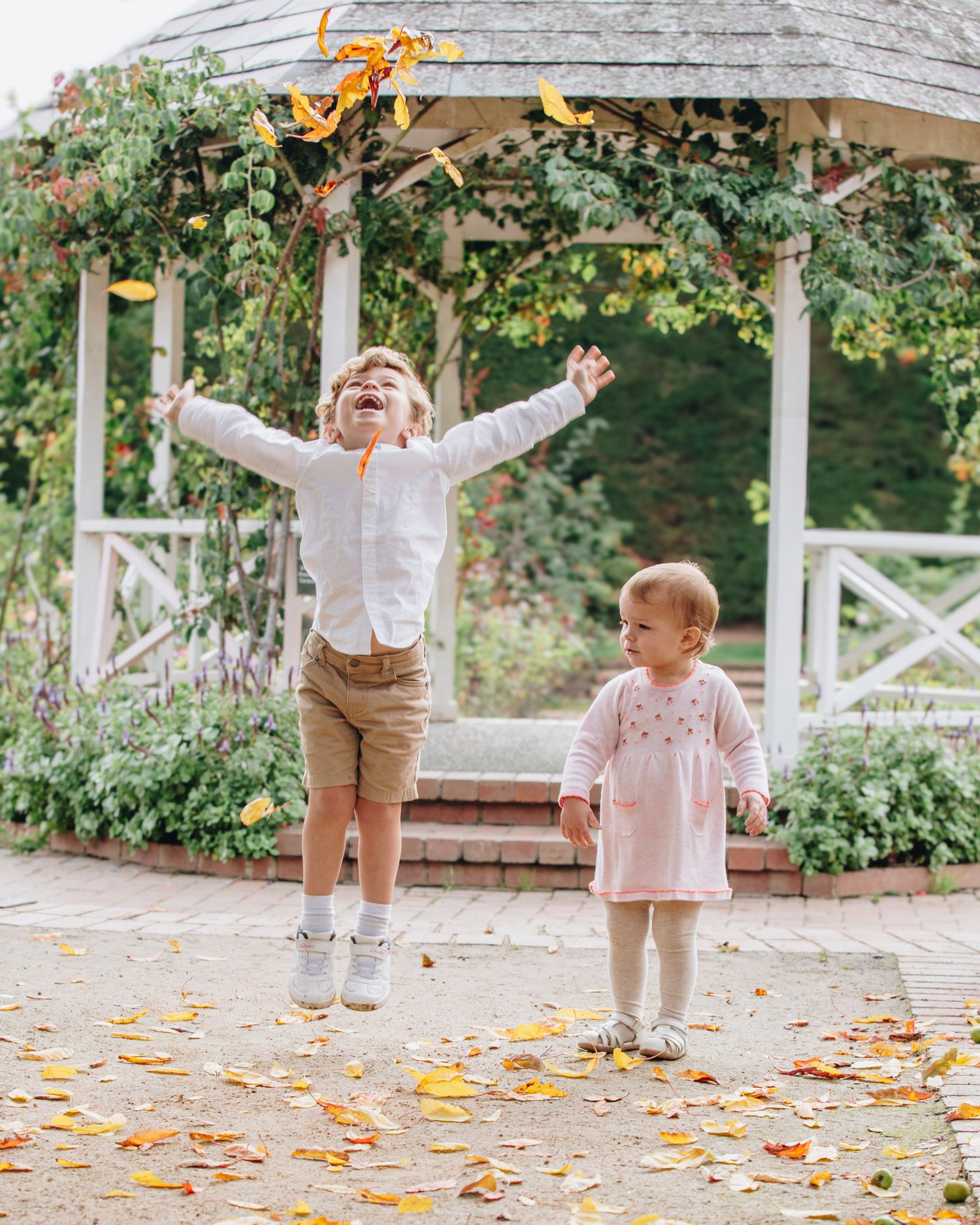 Children playing in garden gazebo with one jumping while surrounded by fallen leaves.