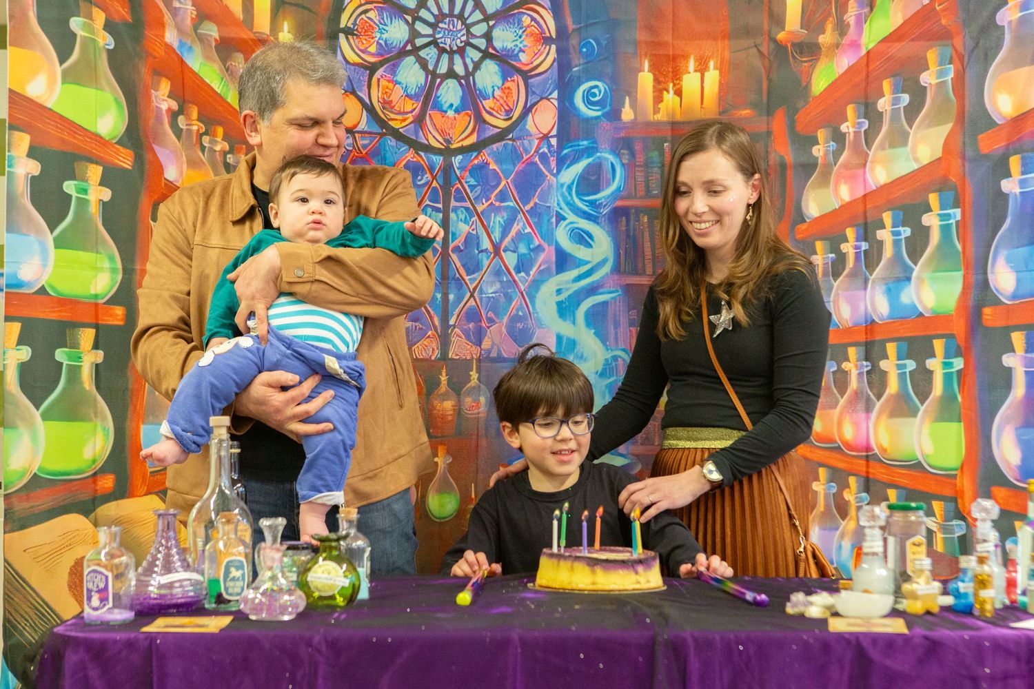 Family celebrating birthday with candles at a colorful potion-themed party backdrop with shelves of bottles.