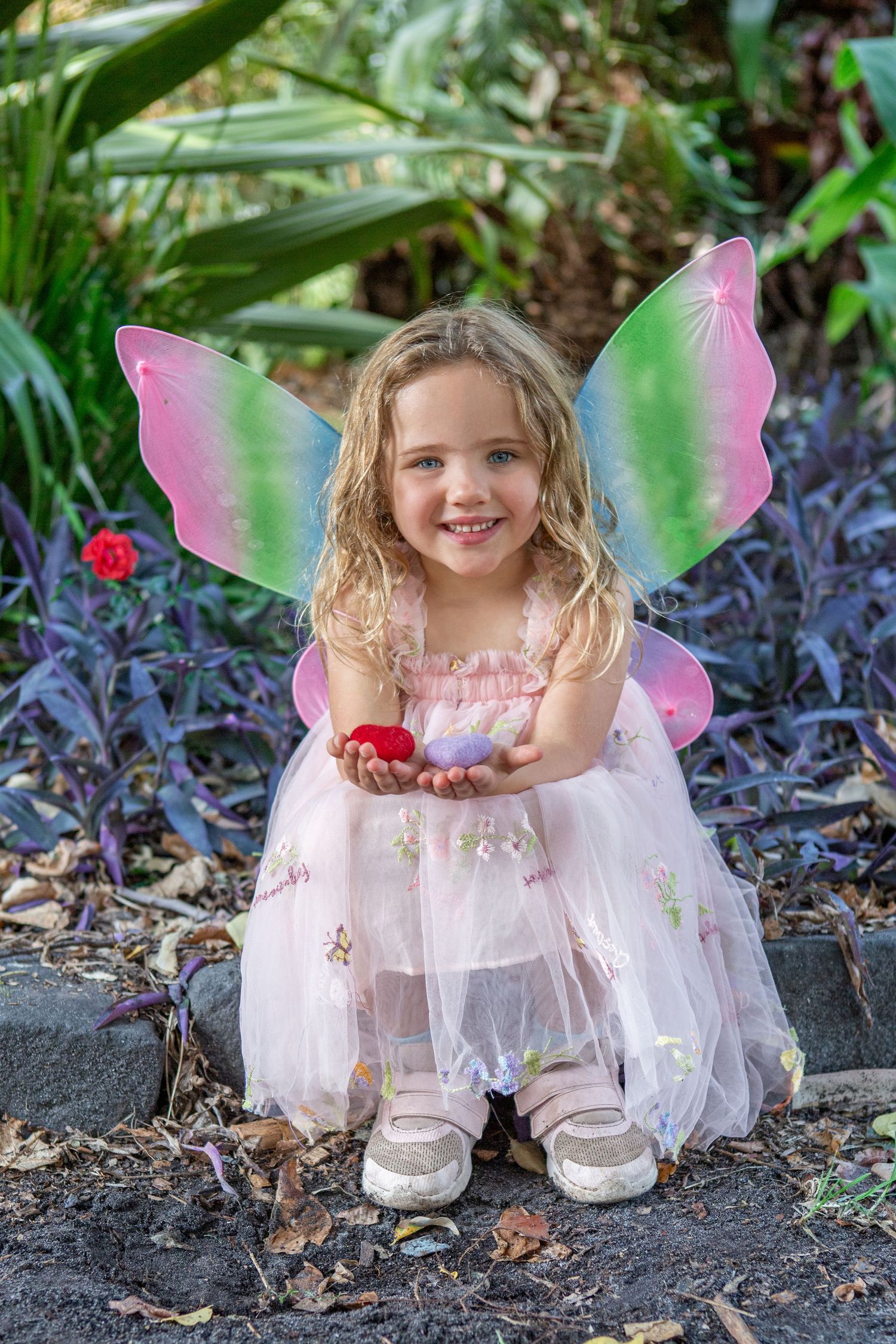 Young girl in pink fairy costume with colorful wings smiles while holding small red heart in garden setting.