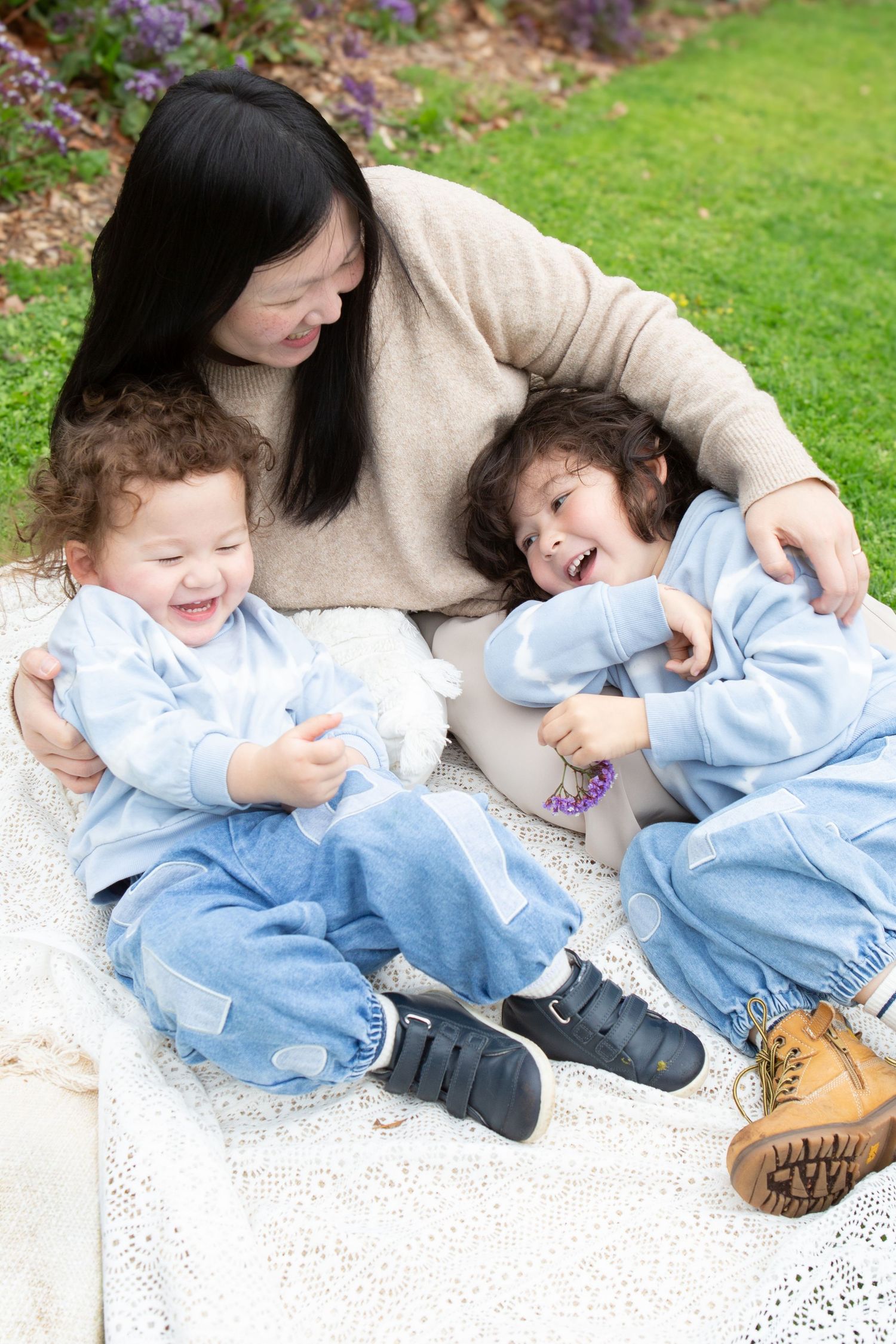 A woman sits on a blanket outdoors laughing with two young children in matching blue outfits on a sunny day.