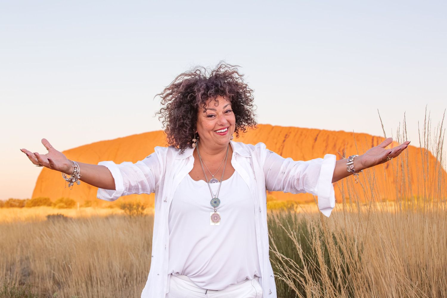 Someone with curly hair spreads their arms wide while wearing white against a sunset desert background.