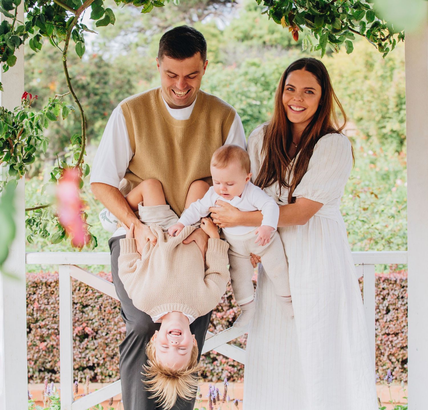 Happy family of four posing under a garden pergola, parents holding baby and playfully flipping toddler upside down.