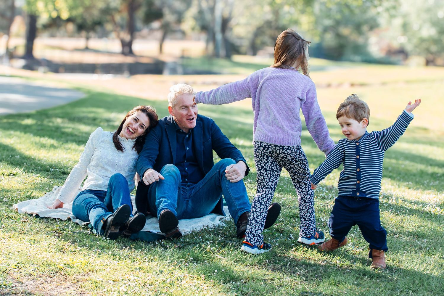 Family enjoying a sunny park day, with grandparents sitting on grass while two young children play nearby.
