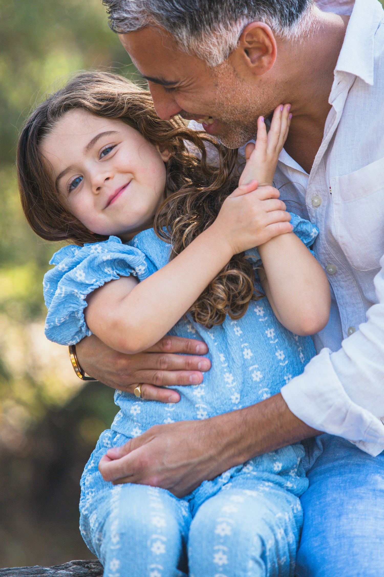 A father tenderly embraces his smiling young daughter in a blue dress outdoors in a green park setting.
