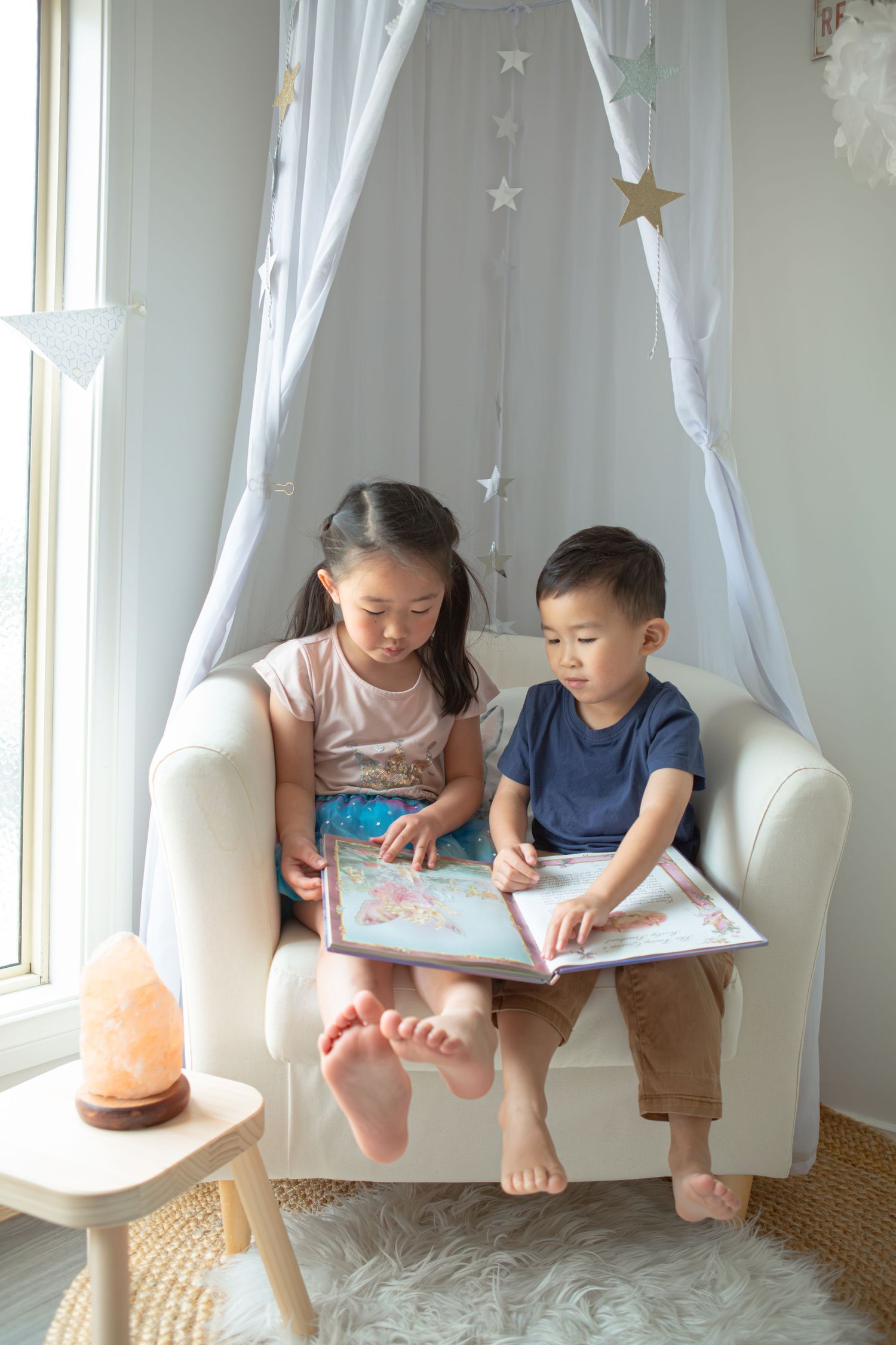 Two young children sitting in a white chair reading a colorful picture book together.