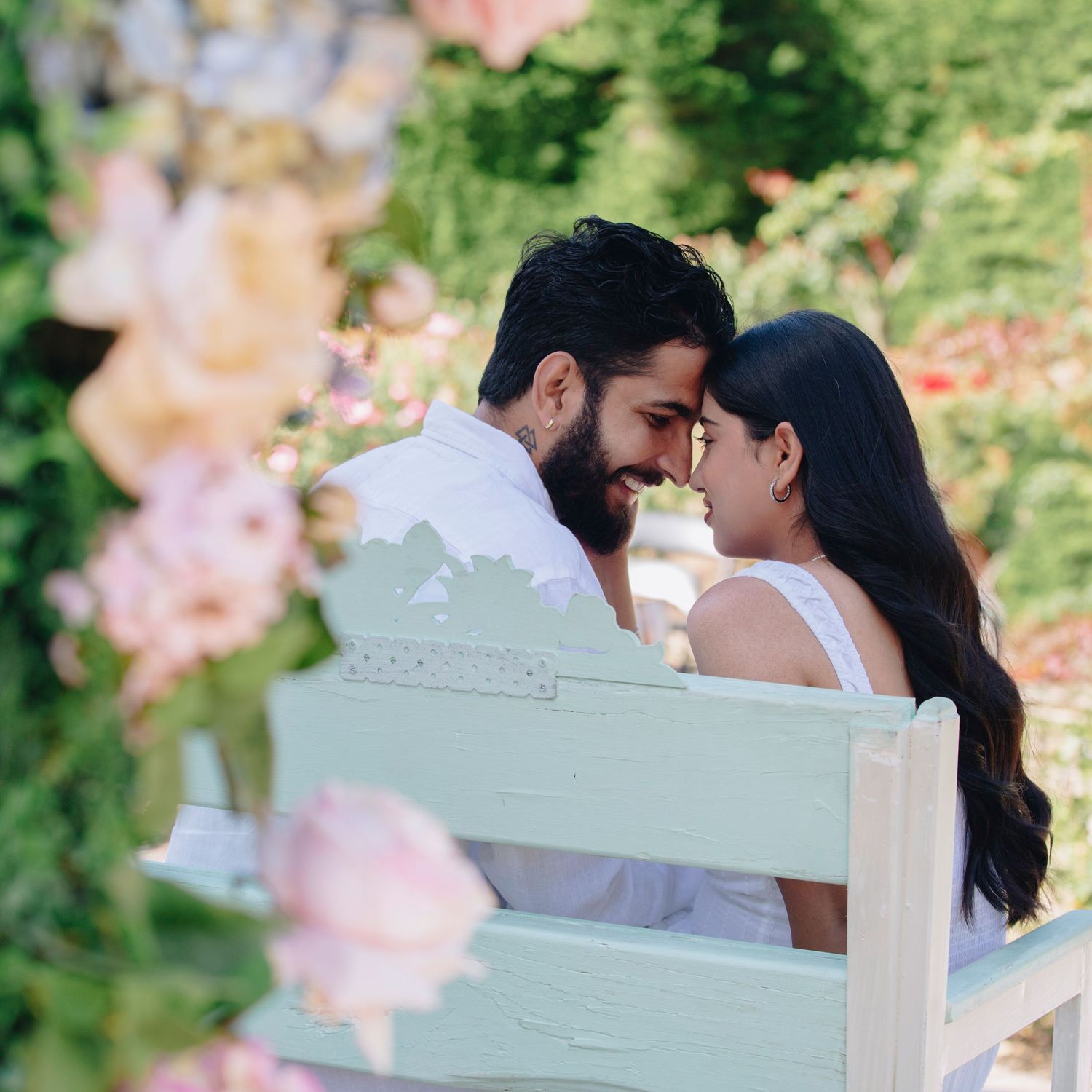 A couple shares a romantic moment on a mint green bench surrounded by lush gardens and soft pink floral arrangements.