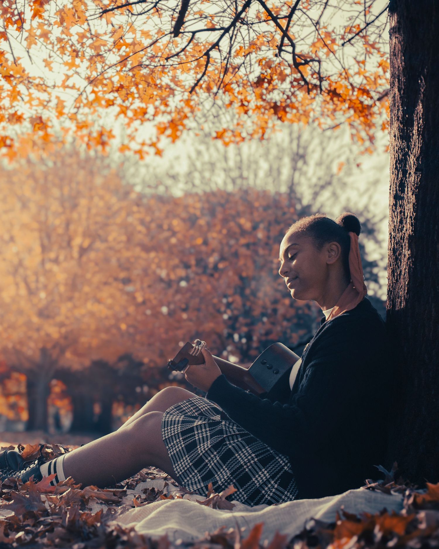 A young woman sits against a tree in a park, reading during golden autumn, wearing a plaid skirt amid fallen leaves.