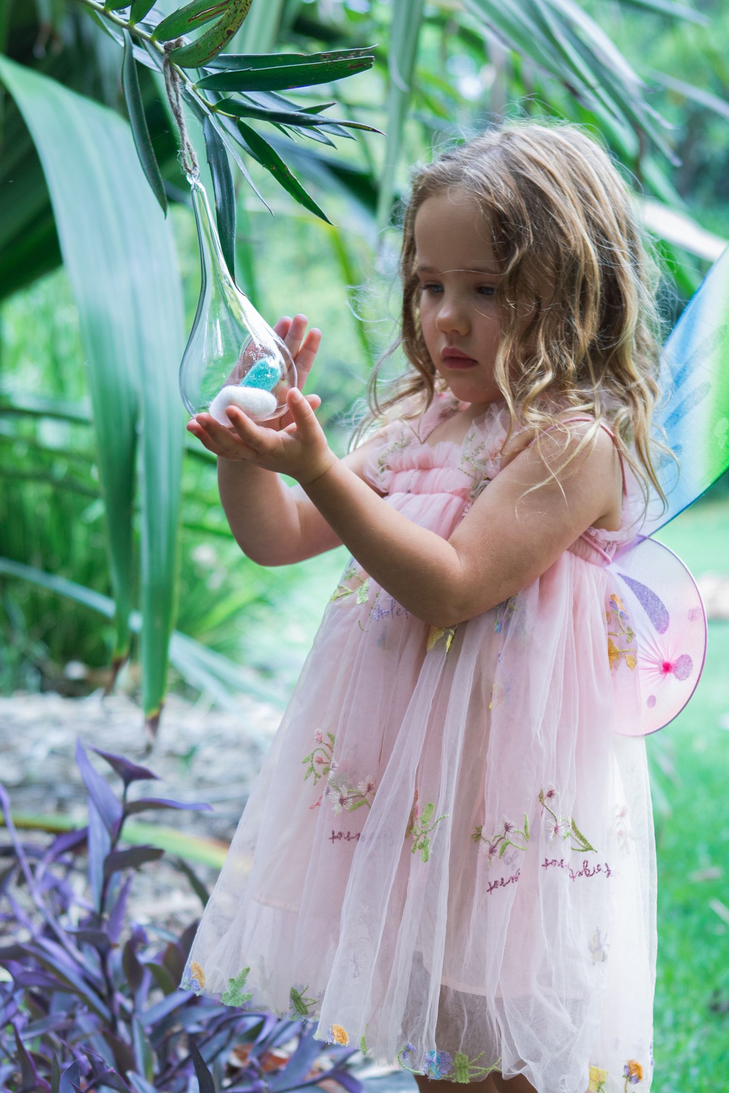 Young girl in pink fairy dress with wings holds a glass ornament among lush tropical garden plants.
