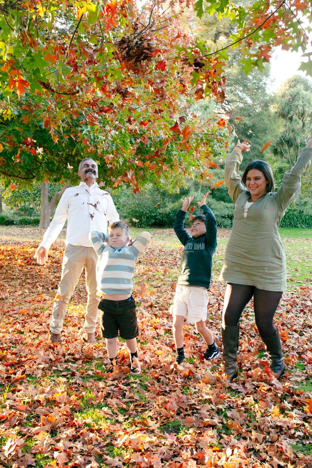 Family of four joyfully tossing autumn leaves in a park surrounded by vibrant red and orange foliage.