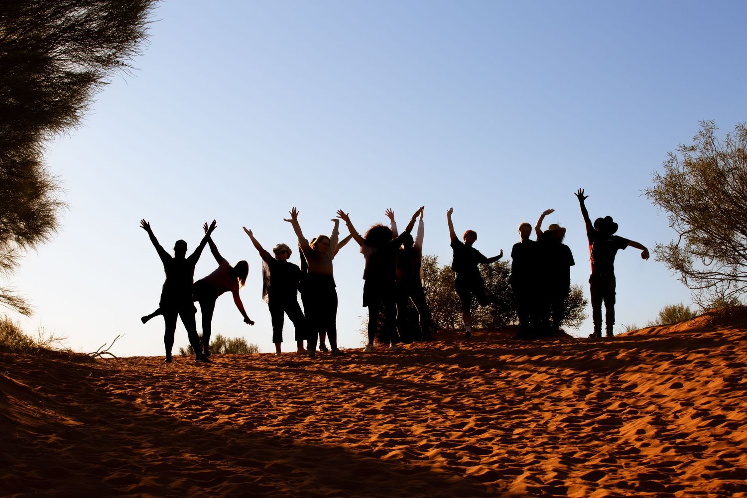 Silhouettes of joyful people jumping and posing on a sandy hill during sunset outdoors.